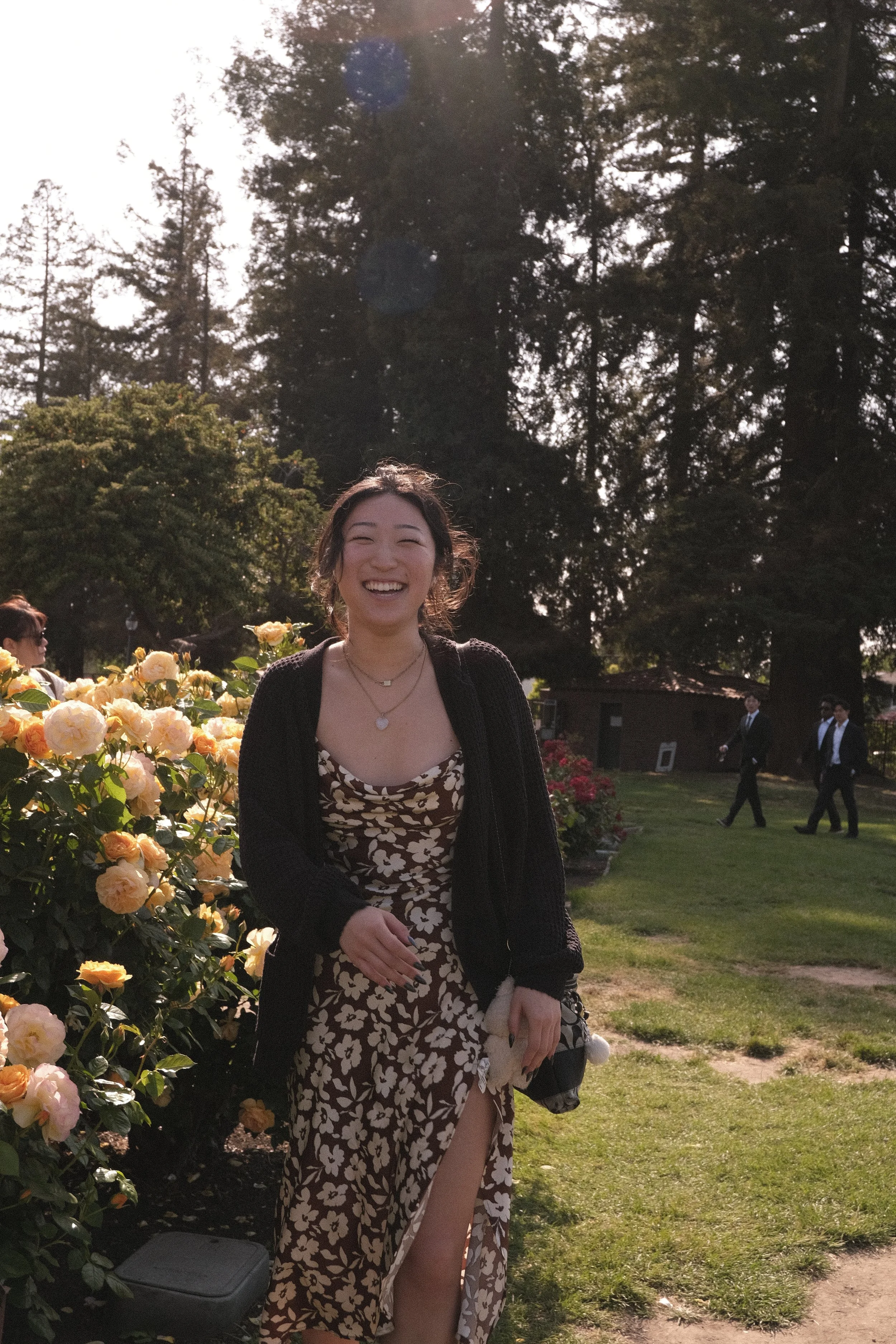 A young woman smiling and standing outdoors in a garden with yellow roses and tall trees in the background. She is wearing a floral dress and multiple necklaces, holding a small stuffed animal and a black purse.
