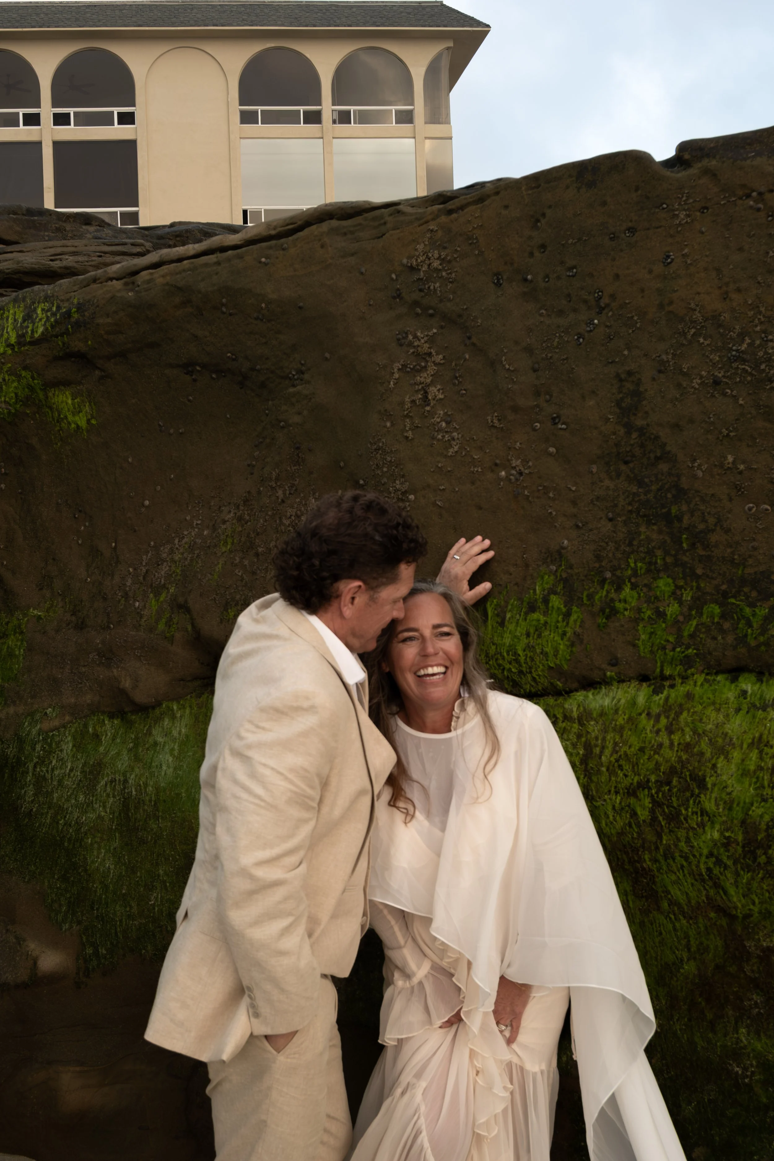 A man and woman smiling and leaning toward each other outdoors, standing against a large moss-covered rock with a building featuring arched windows behind them.