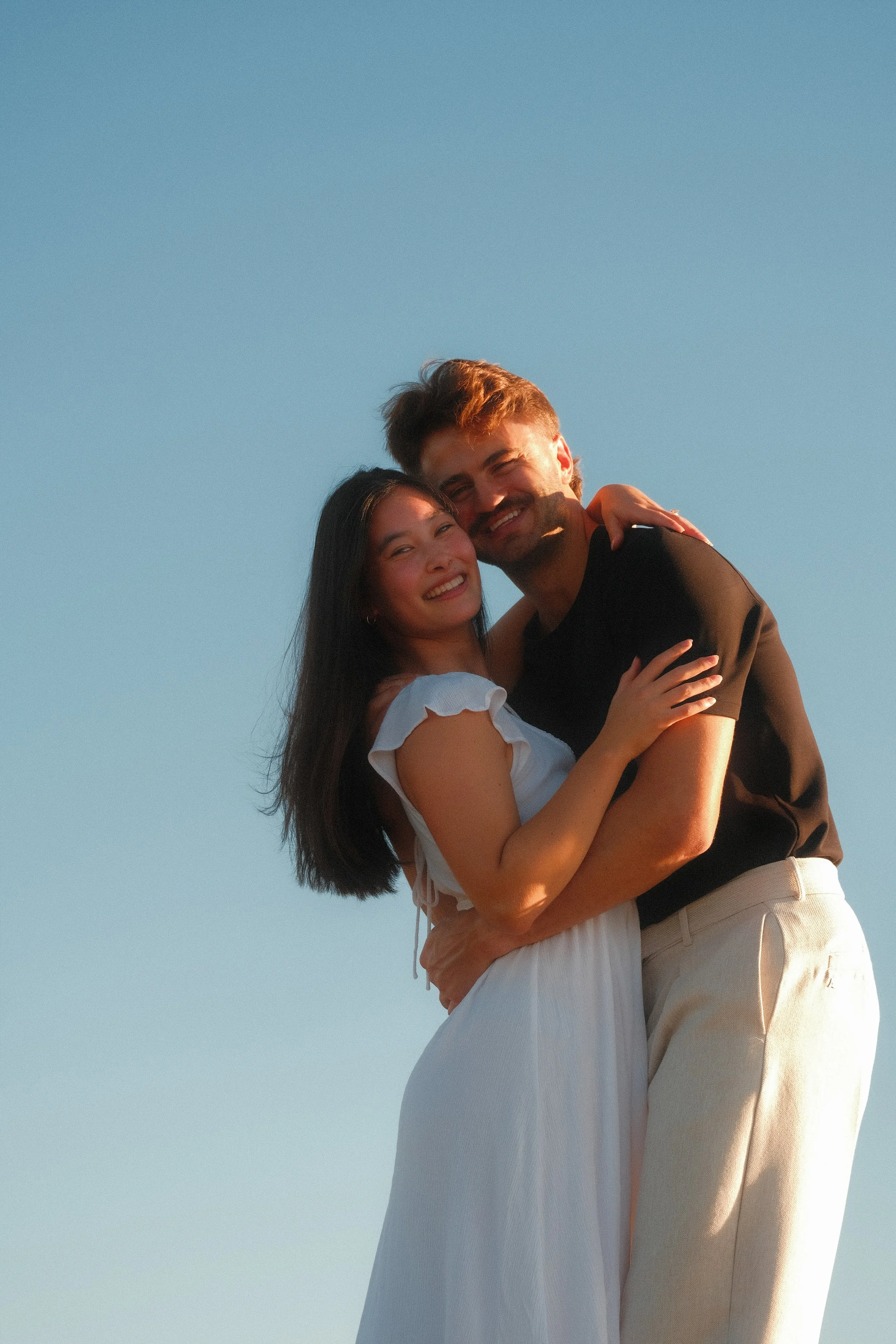 A couple is embracing outdoors against a clear blue sky, smiling, with the woman wearing a white dress and the man in a black shirt and beige pants.