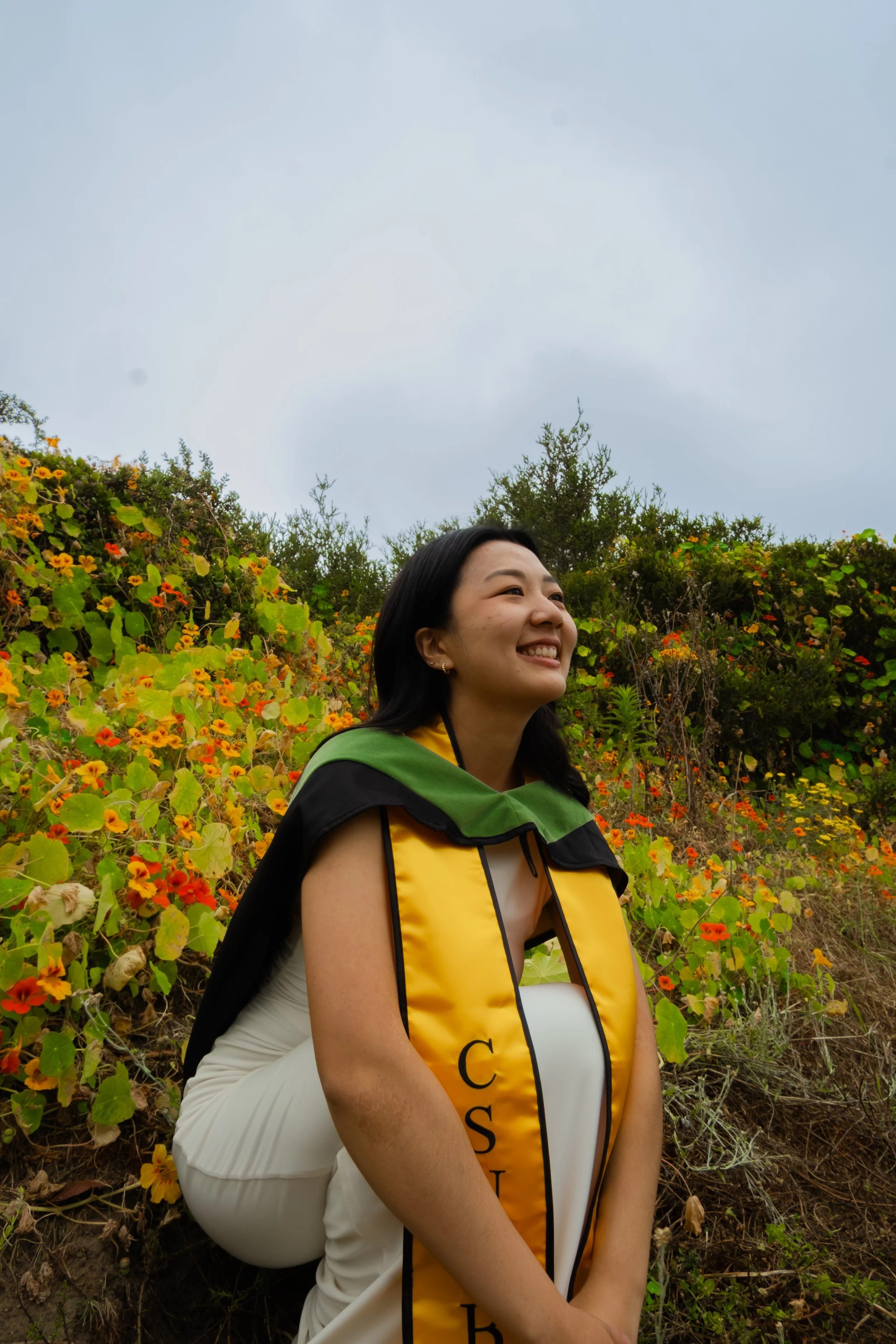 A woman smiling outdoors, wearing a graduation cap and gown with a yellow stole, standing in front of bushes with orange and yellow flowers, under a cloudy sky.