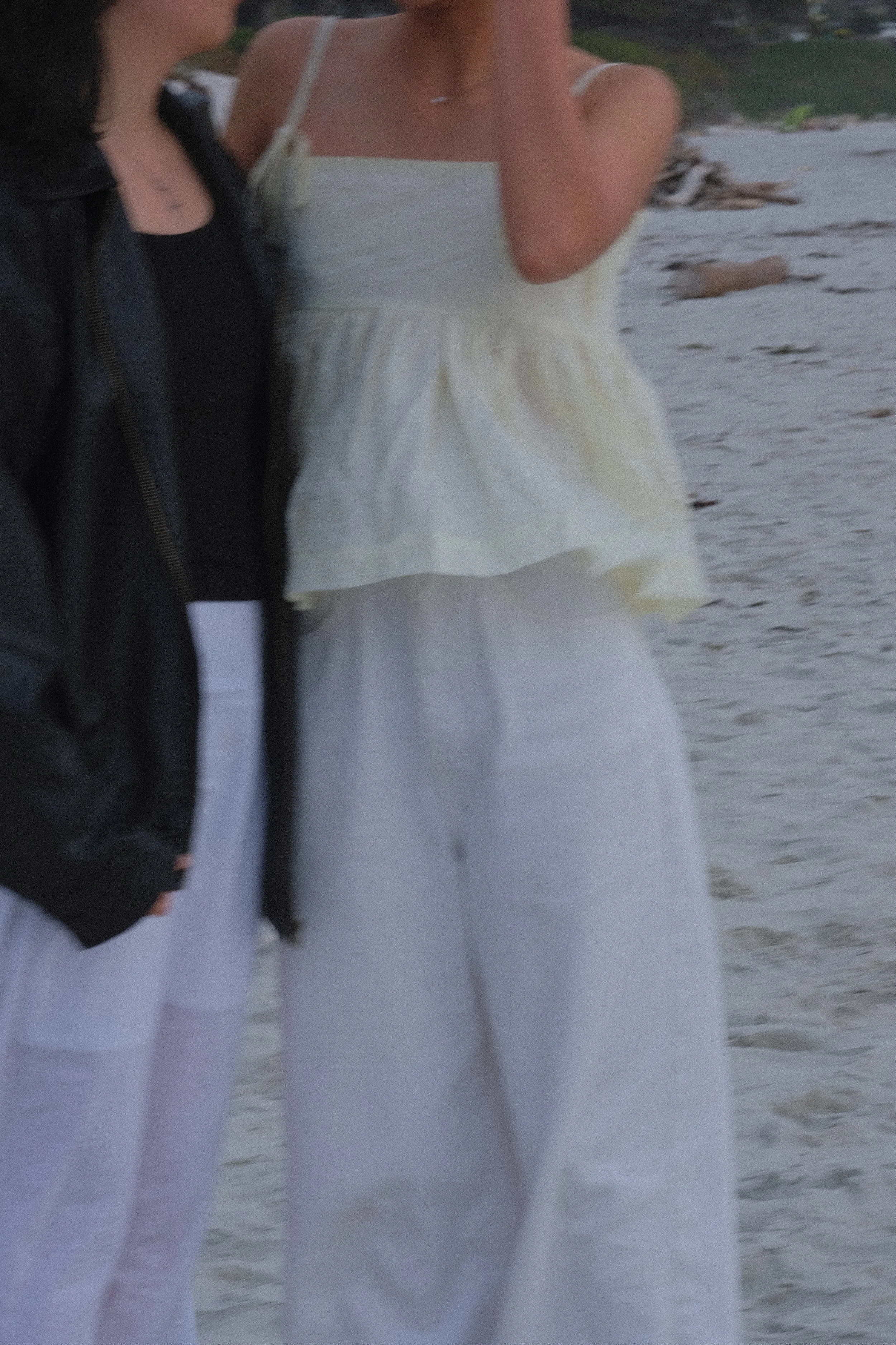 Two women standing on a beach, one wearing a white sundress and the other wearing a black jacket and black top.