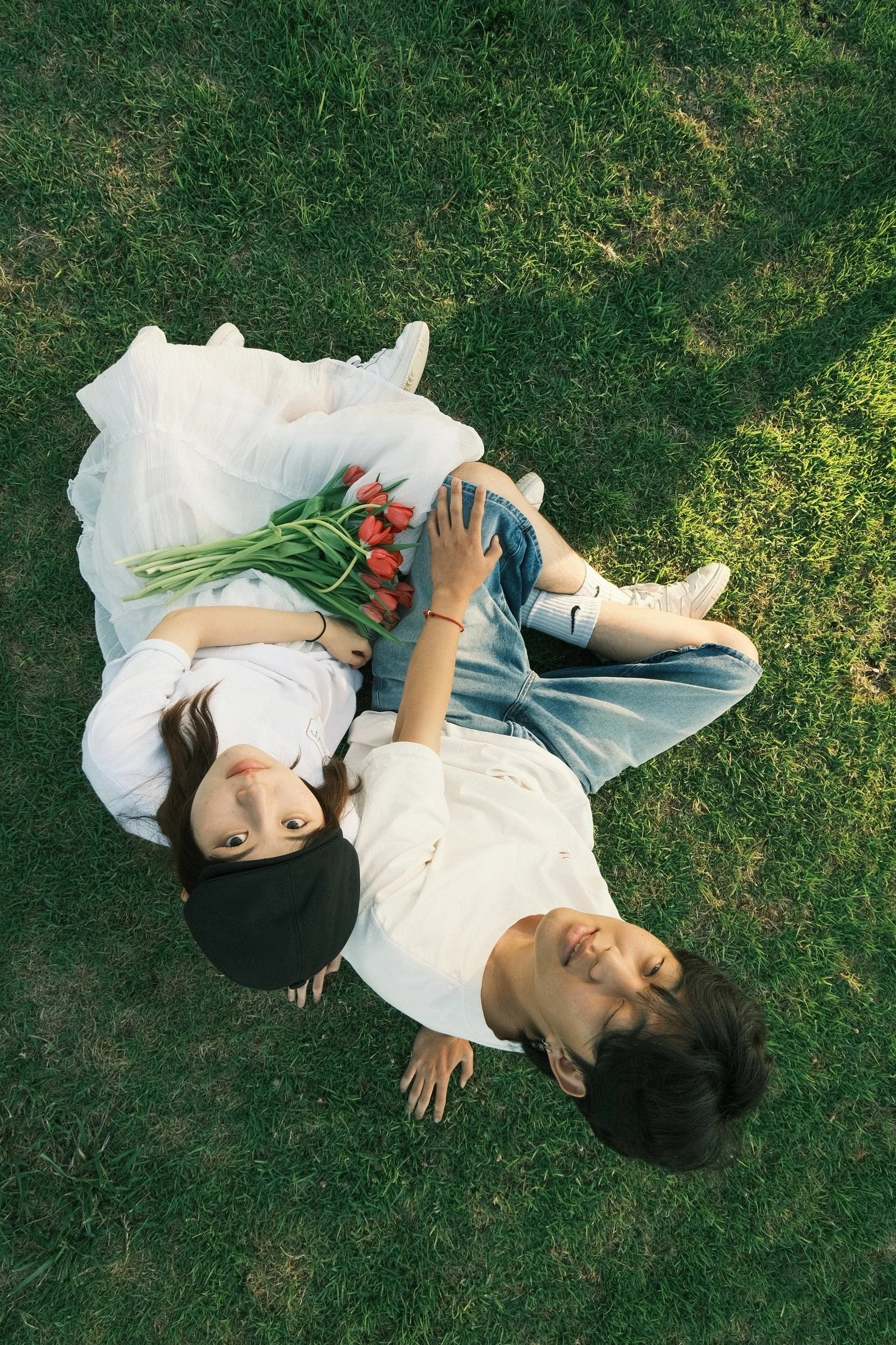 Two young women and a young man are lying on green grass, one of the women is holding a bouquet of red tulips, all three are relaxed and looking up at the camera.