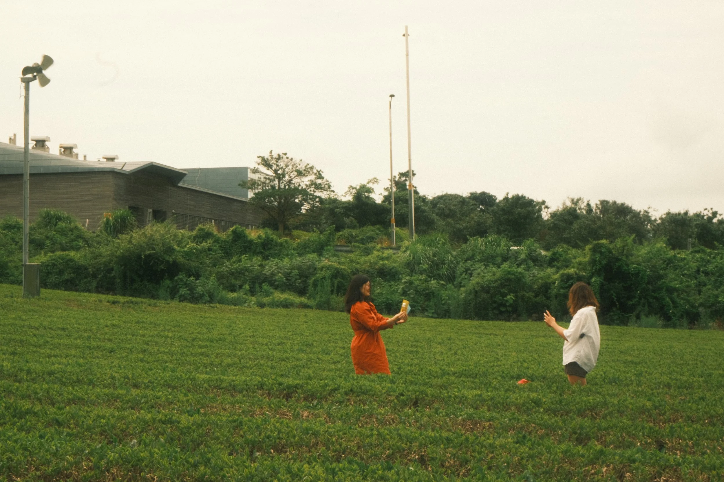 Two women standing on a grassy area, one wearing an orange jacket and the other wearing a white jacket, engaging in conversation.