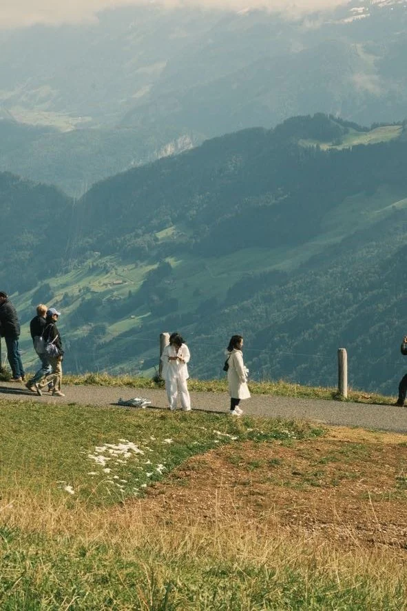 People hiking on a grassy trail overlooking a valley with mountains in the background.