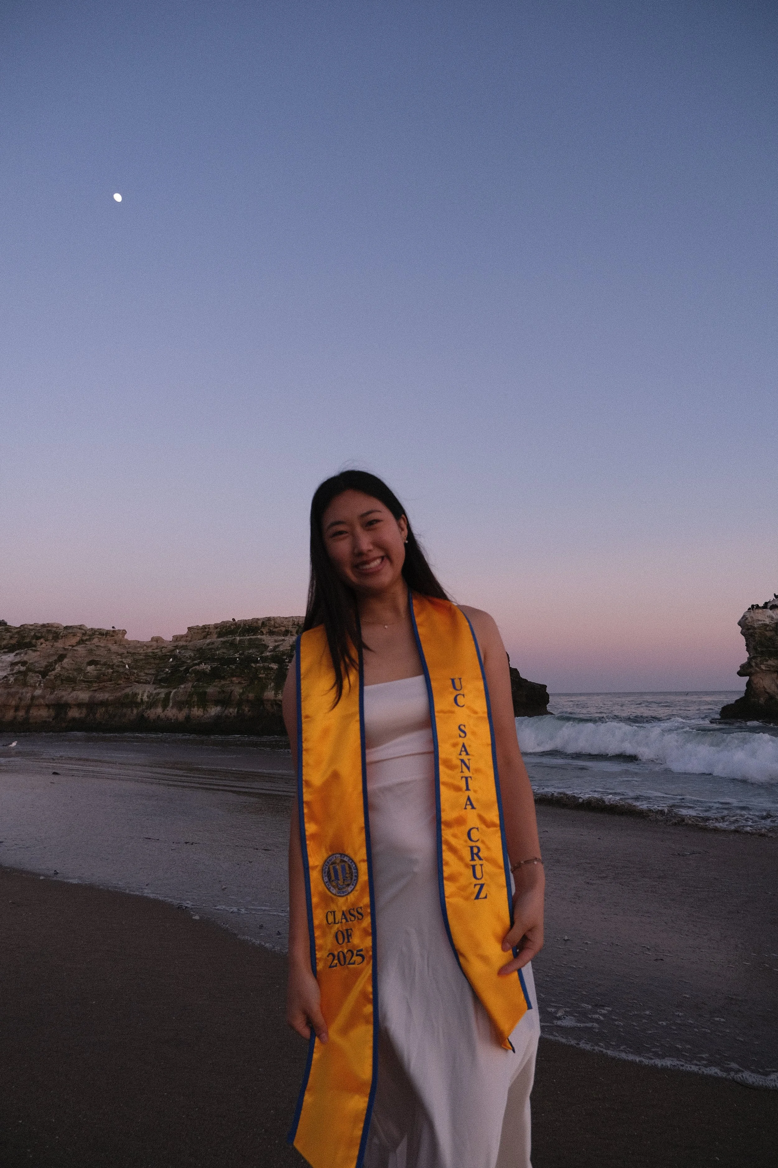 Young woman smiling on a beach at sunset, wearing a yellow sash with blue border that reads 'U.C.S. Santa Cruz,' white dress, and a necklace.