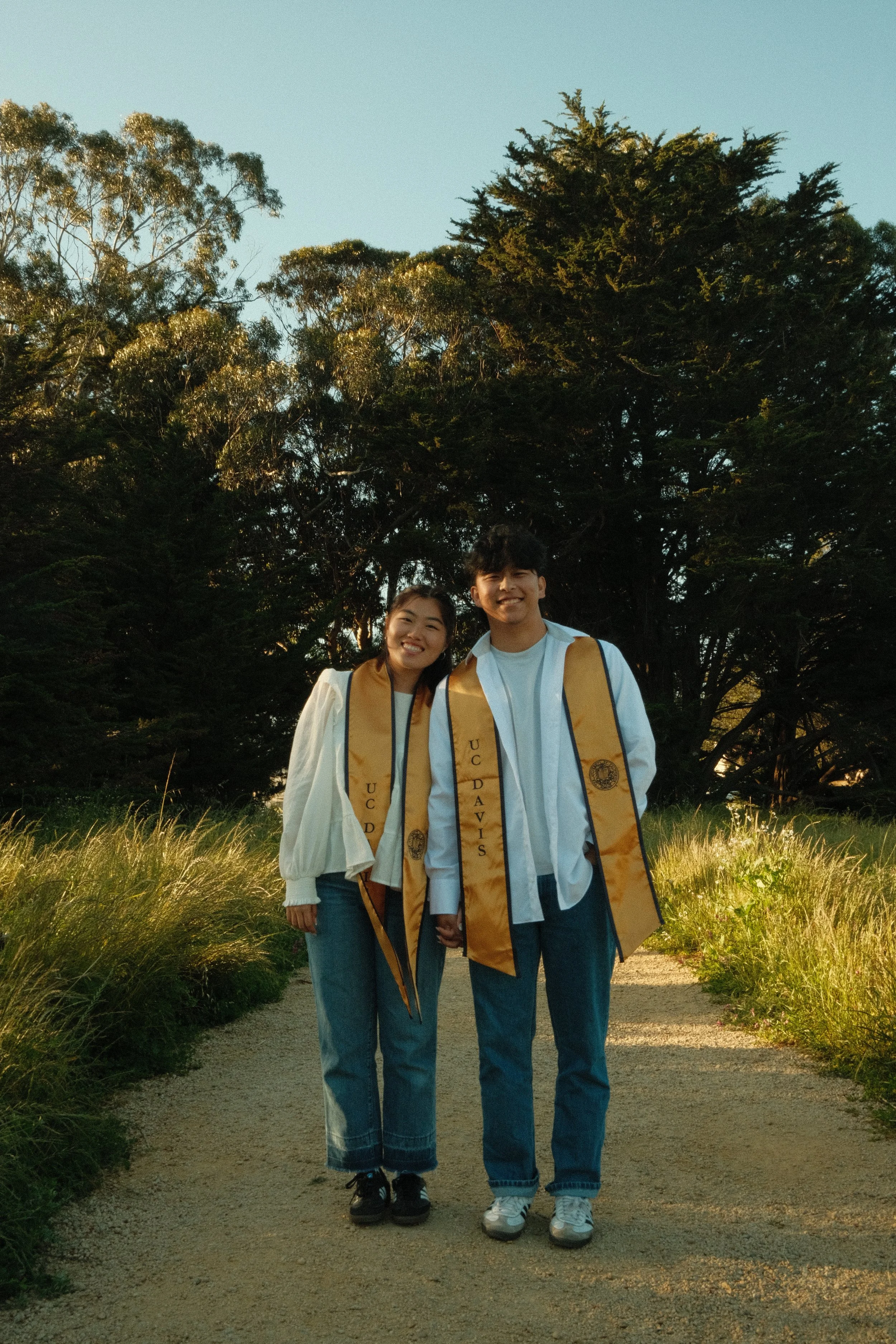 Two smiling young adults, a woman and a man, wearing graduation gowns and stoles, standing on a dirt path surrounded by green plants with tall trees and a clear blue sky in the background during daytime.