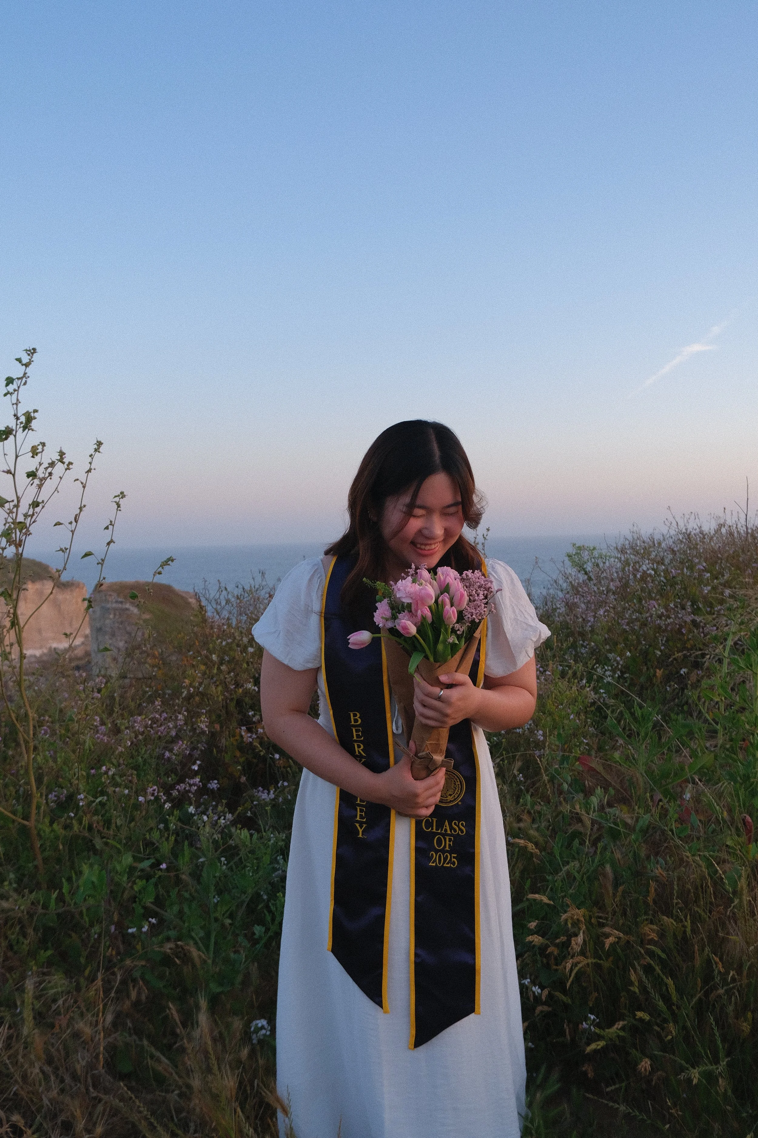 A young woman in a white dress, wearing a black and gold graduation stole that reads "BEREY CLASS OF 2025," is holding a bouquet of pink and purple flowers and smiling while standing outdoors at sunset amid tall grass and plants with a distant view of hills or cliffs.