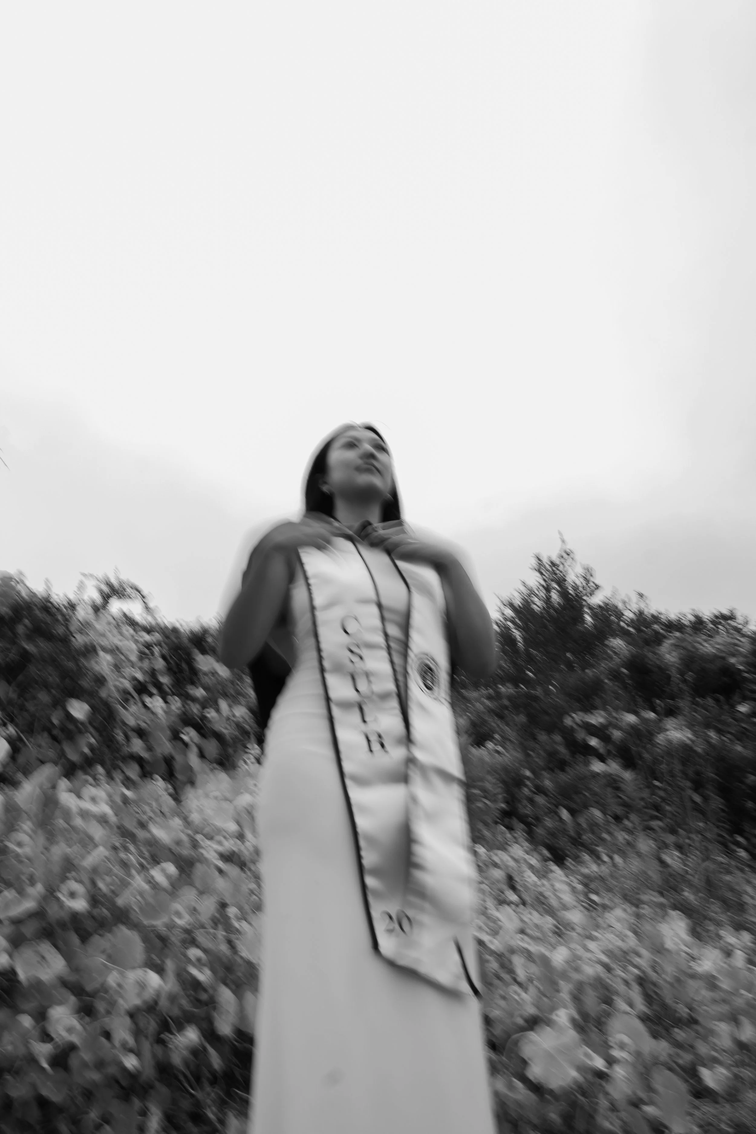 Black and white photo of a woman in outdoor setting, wearing a jacket, standing among bushes and trees, with an overcast sky above.