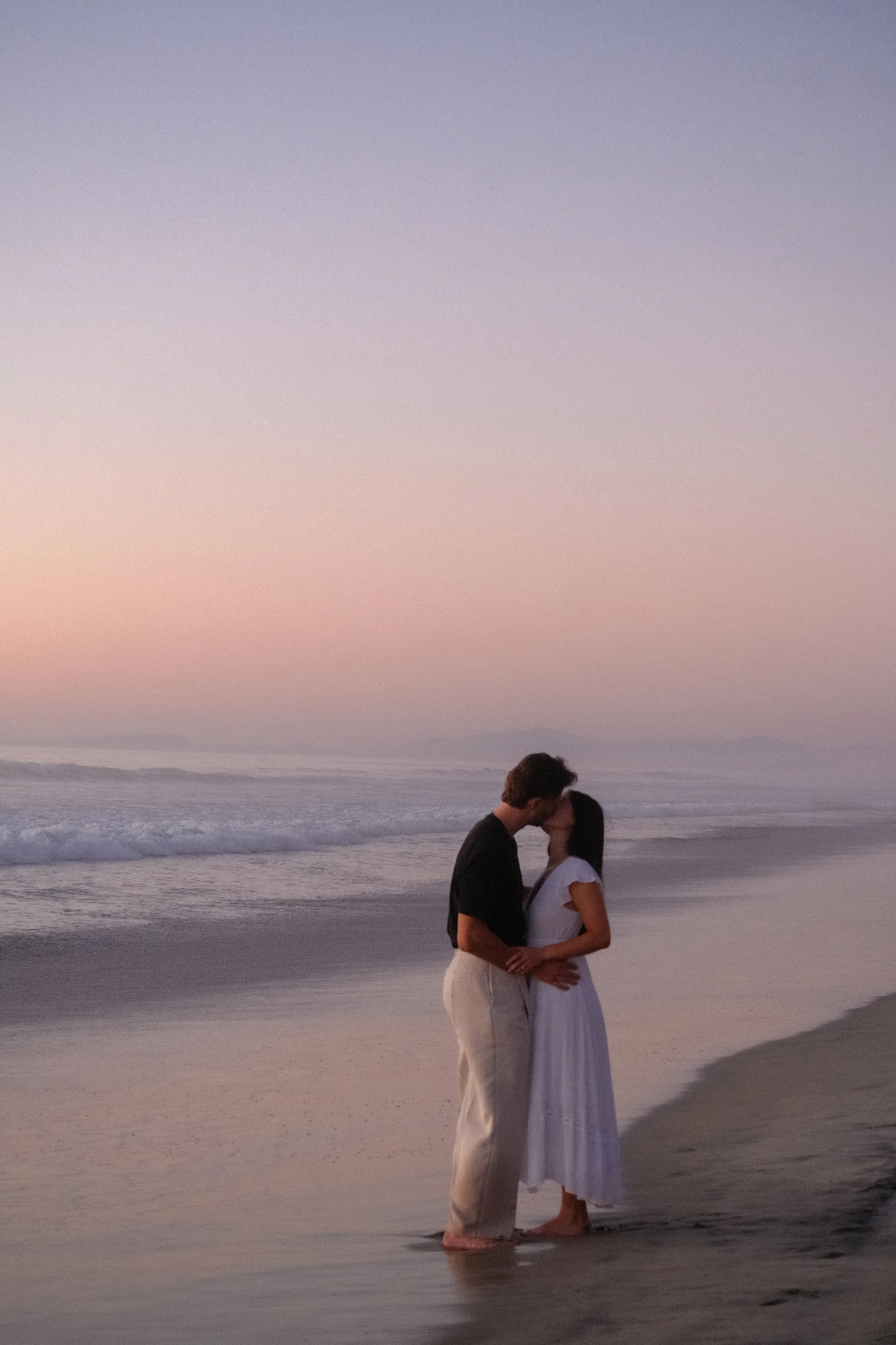 A couple sharing a kiss on the beach during sunset, standing close with the ocean and sky in the background.
