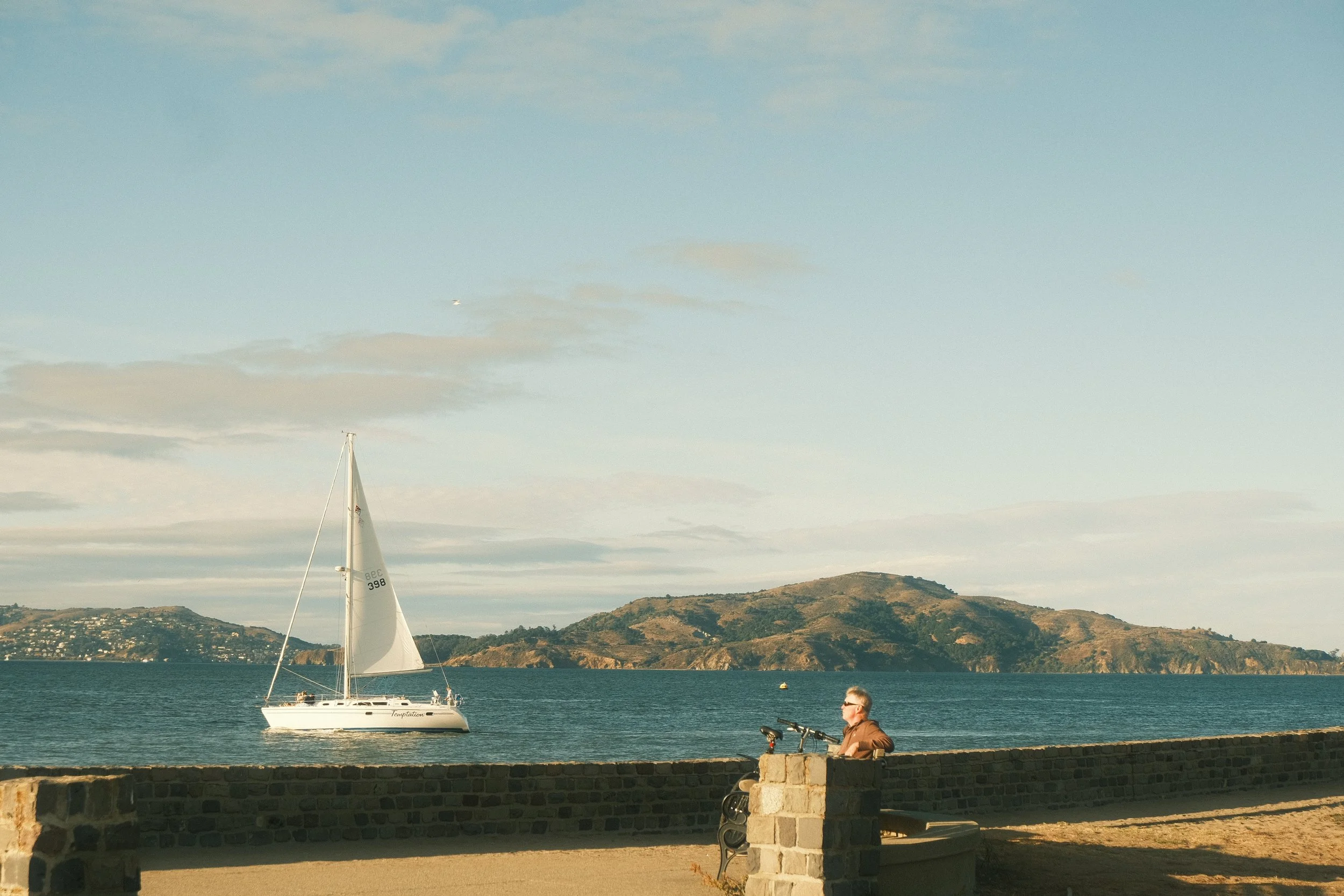 A person with gray hair and sunglasses sitting on a stone wall near the water, with a sailboat named 'Tentation' sailing in the bay, hills in the background, and partly cloudy sky.