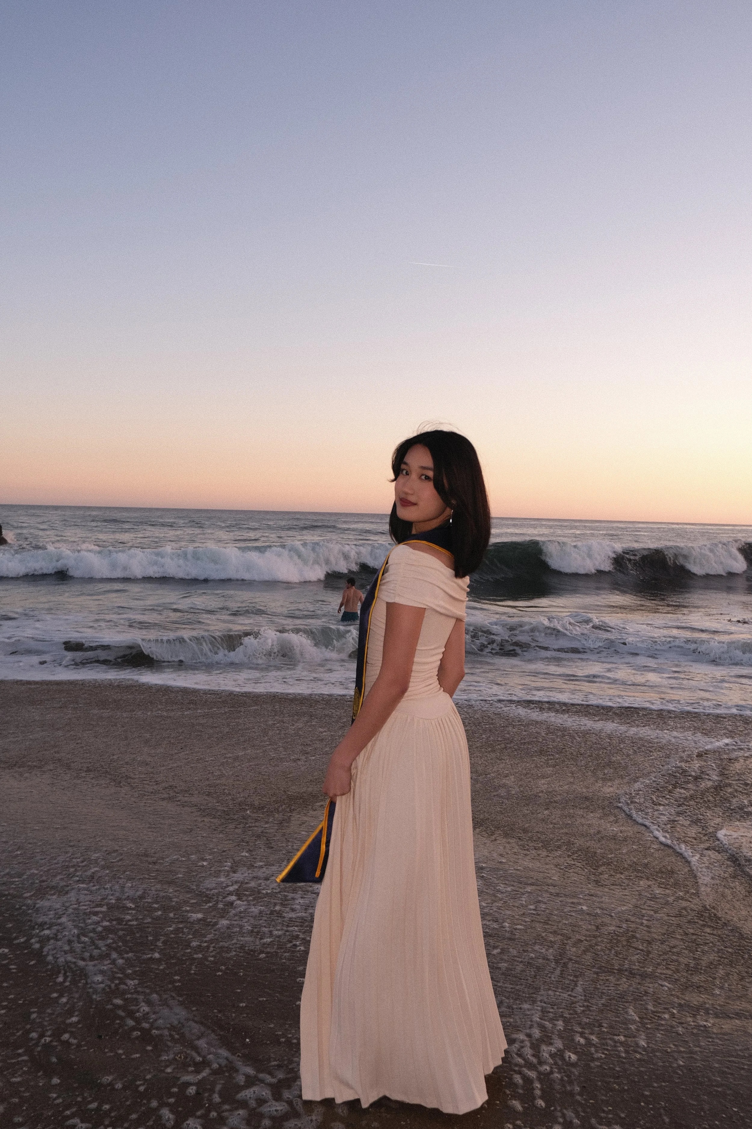 Young woman in a beige pleated dress standing on wet sand at the beach during sunset, with gentle waves in the background and a clear pastel-colored sky.