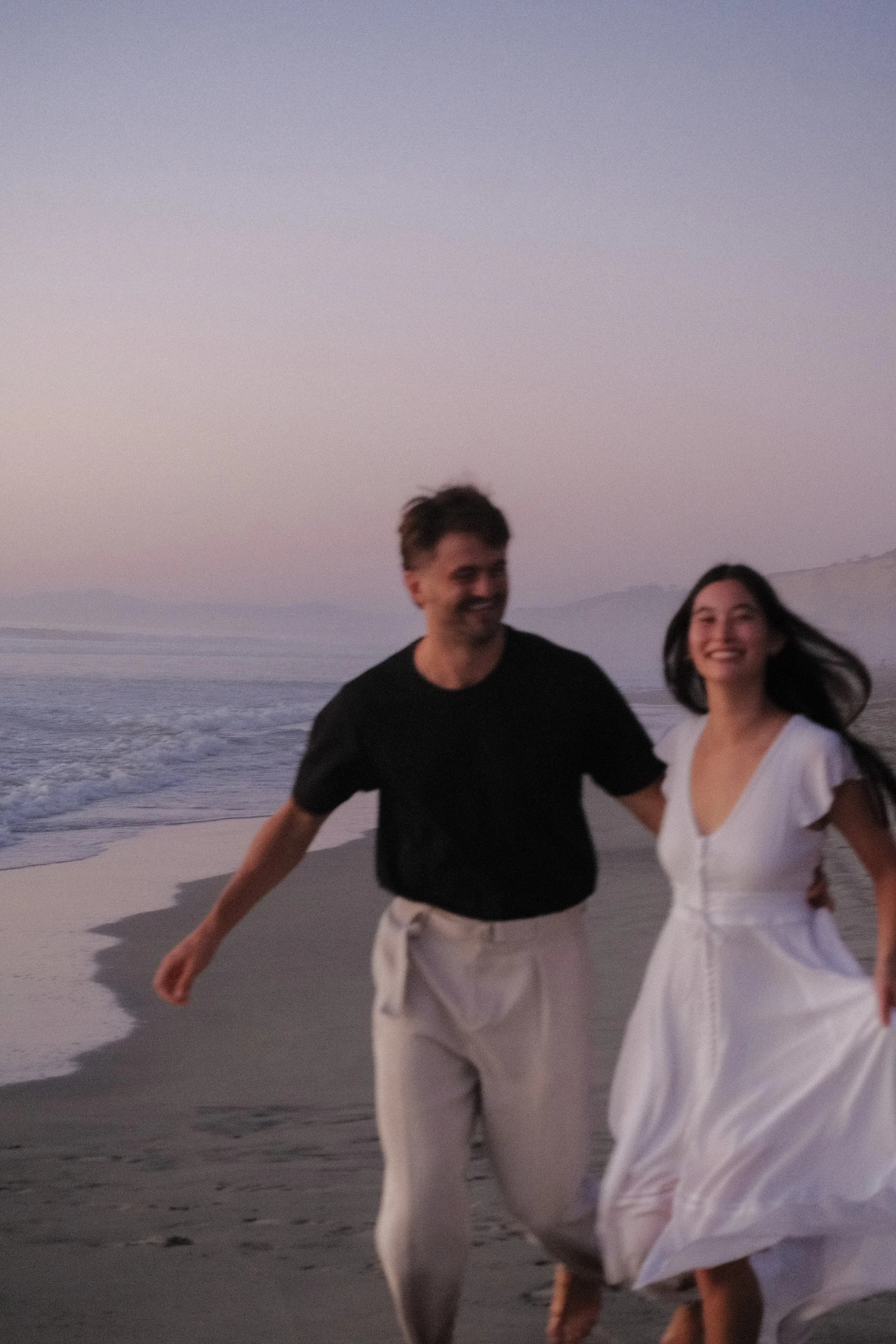 A man and woman are running on a beach during sunset or sunrise, with the ocean waves behind them. The man is wearing a black t-shirt and beige pants, and the woman is wearing a white dress. Both are smiling and appear to be enjoying the moment.