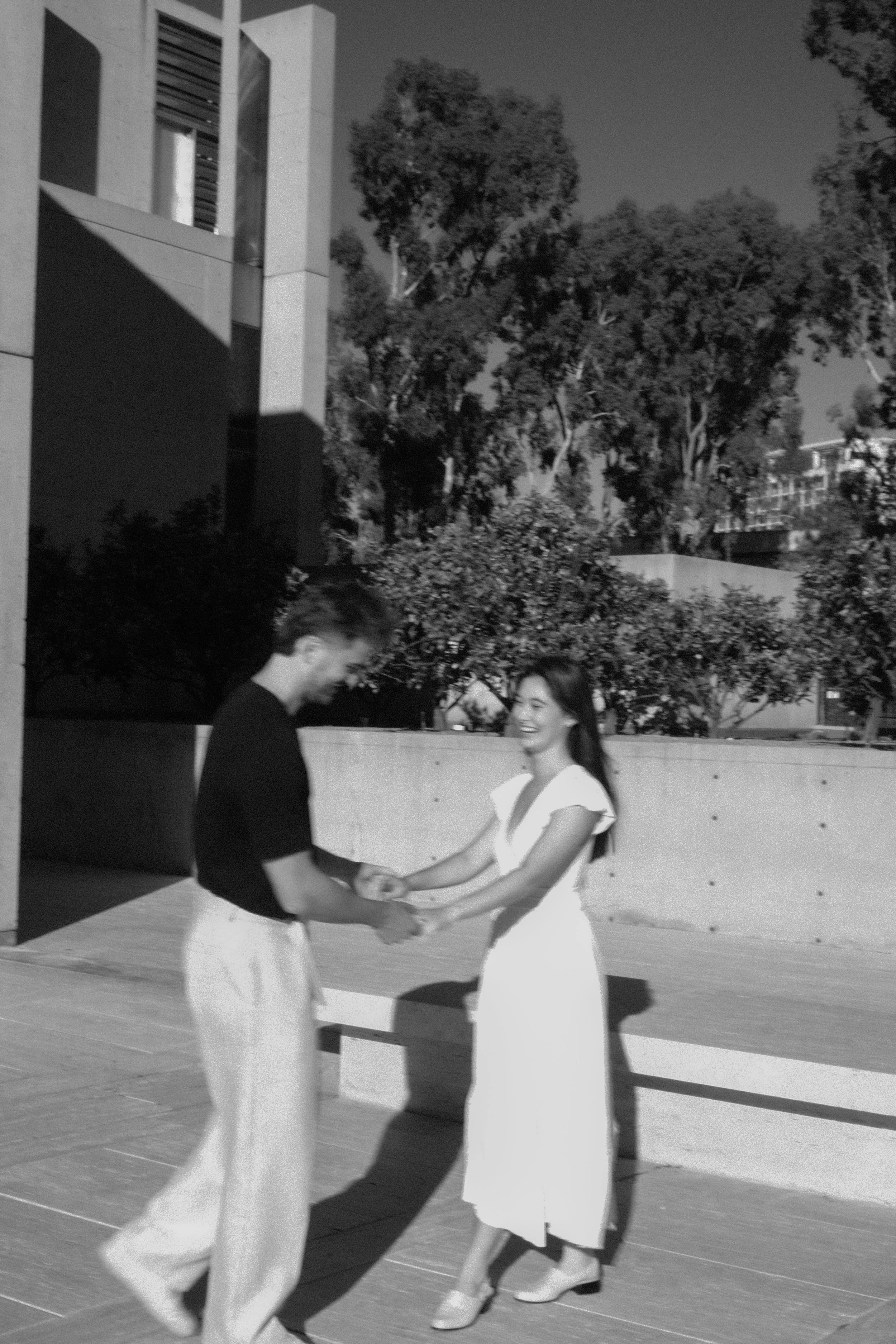 A black and white photo of a young man and woman dancing and holding hands outdoors in front of trees and a building. They are smiling and appear to be enjoying the moment.