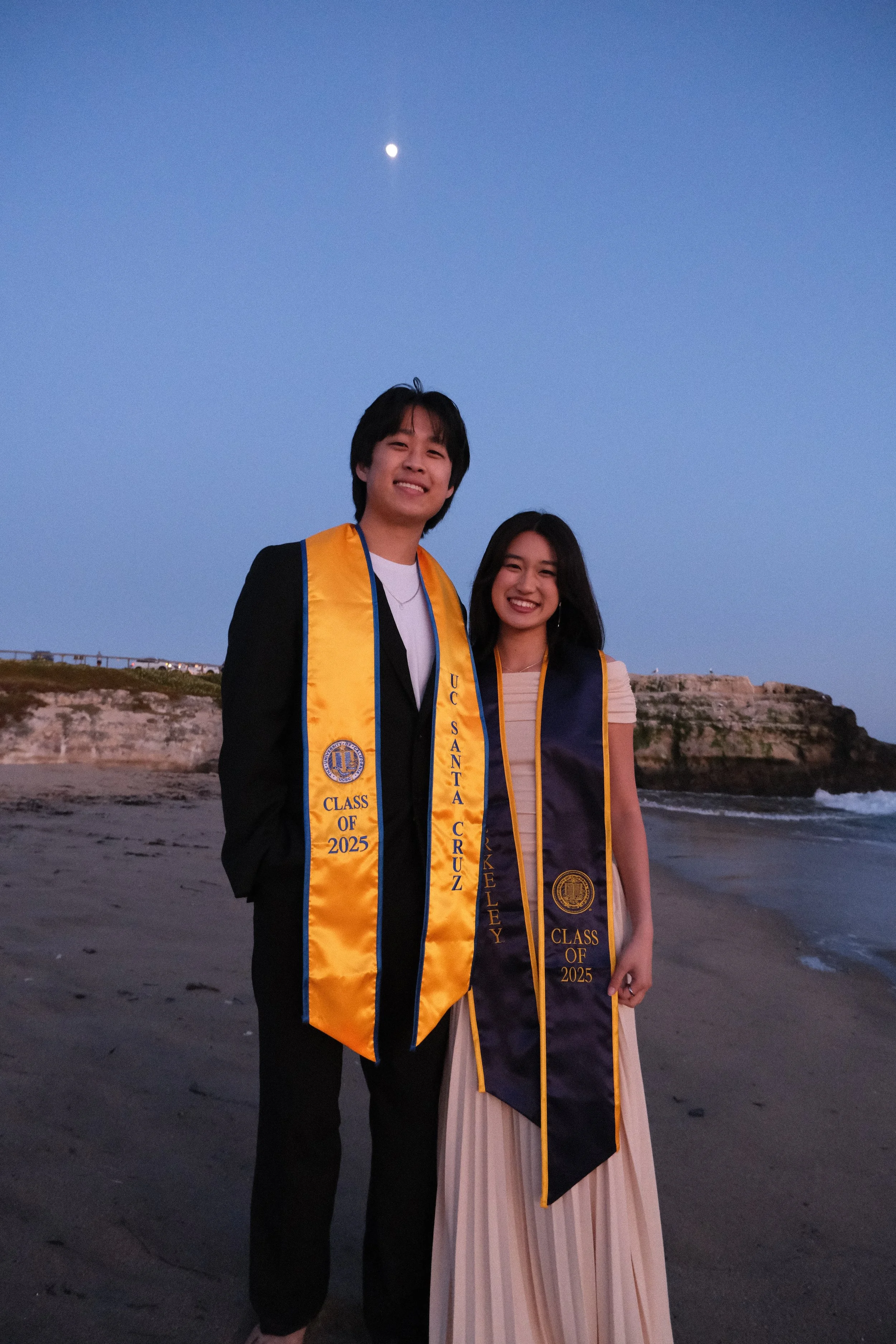 Two young adults, a man and a woman, dressed in graduation gowns with medals and sashes, standing on a beach during dusk, with the moon visible in the sky behind them.