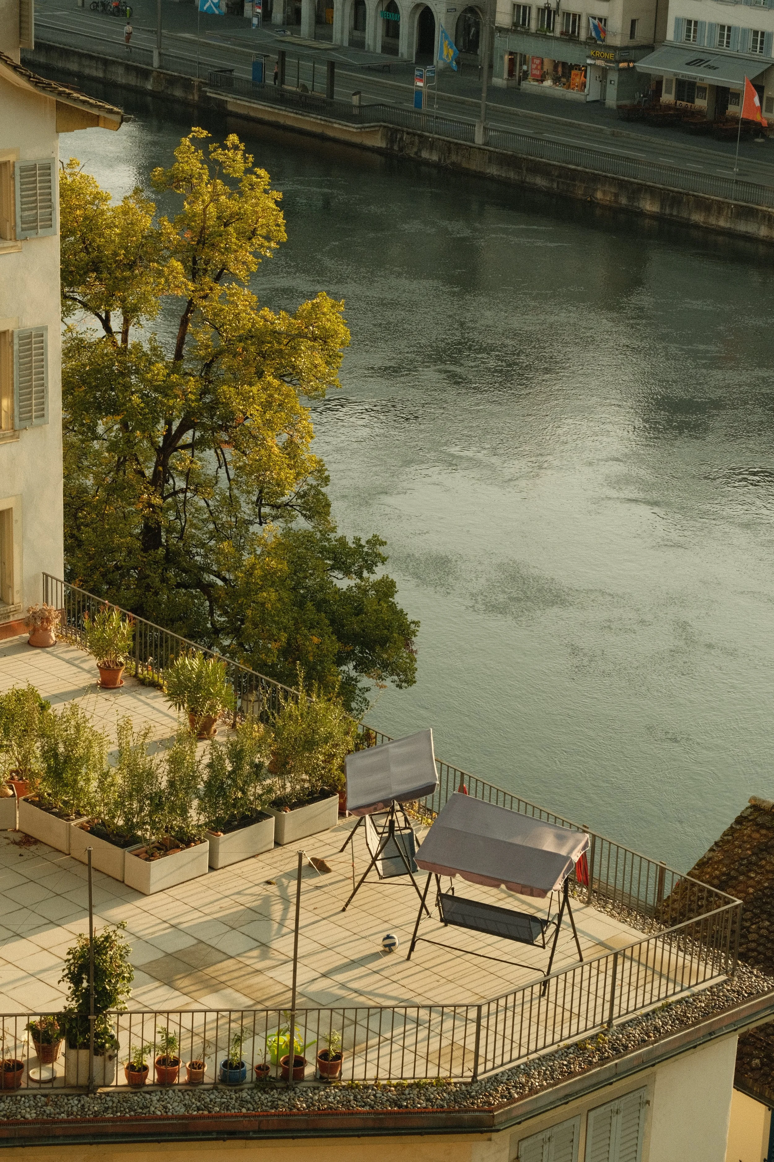 A view of a city rooftop terrace overlooking a river, with potted plants, chairs, and umbrellas, and an urban street with buildings across the water.