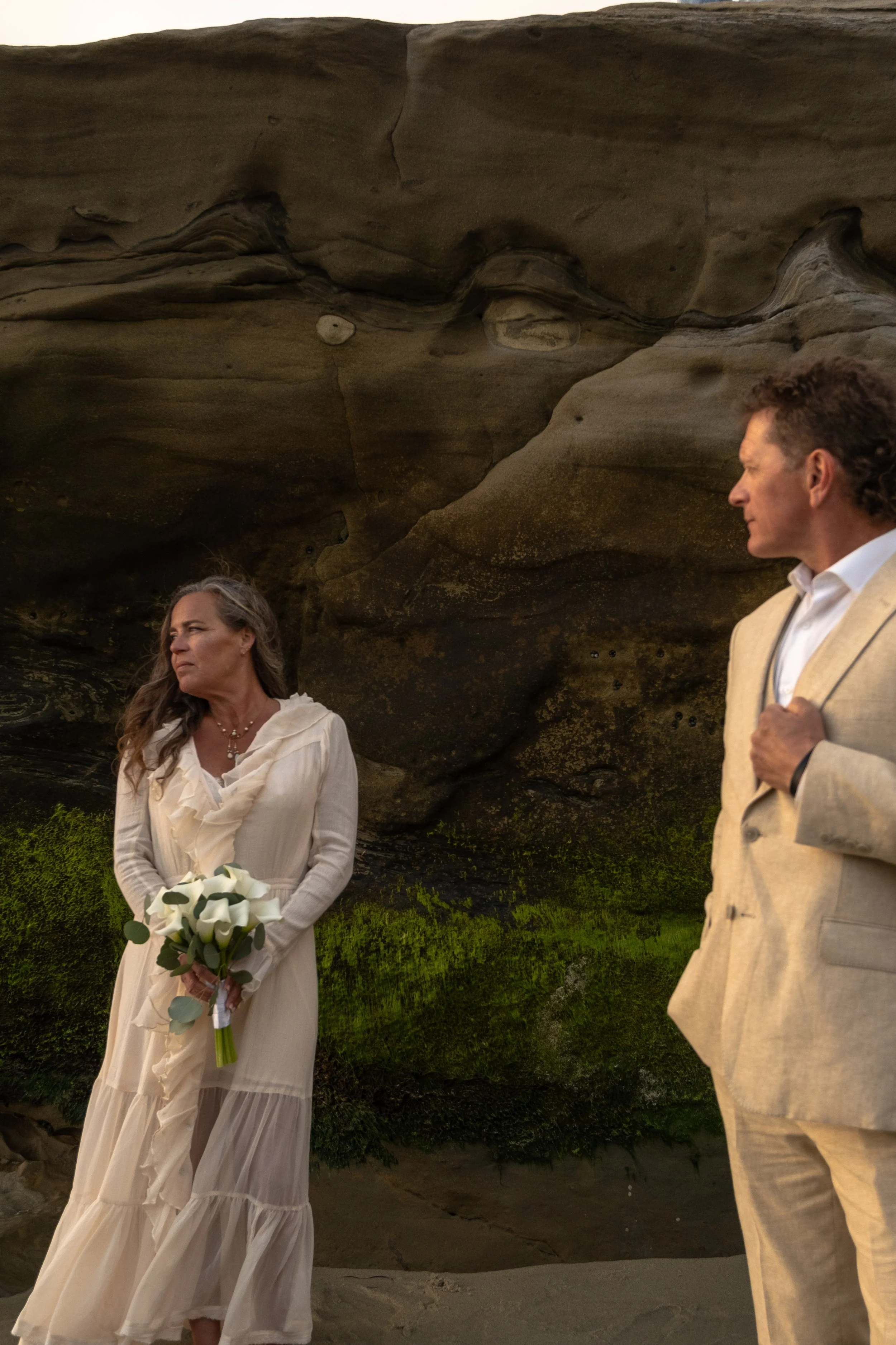 A woman in a white dress holding a bouquet of calla lilies standing outdoors in front of a rocky formation, with a man in a beige blazer standing nearby at sunset.