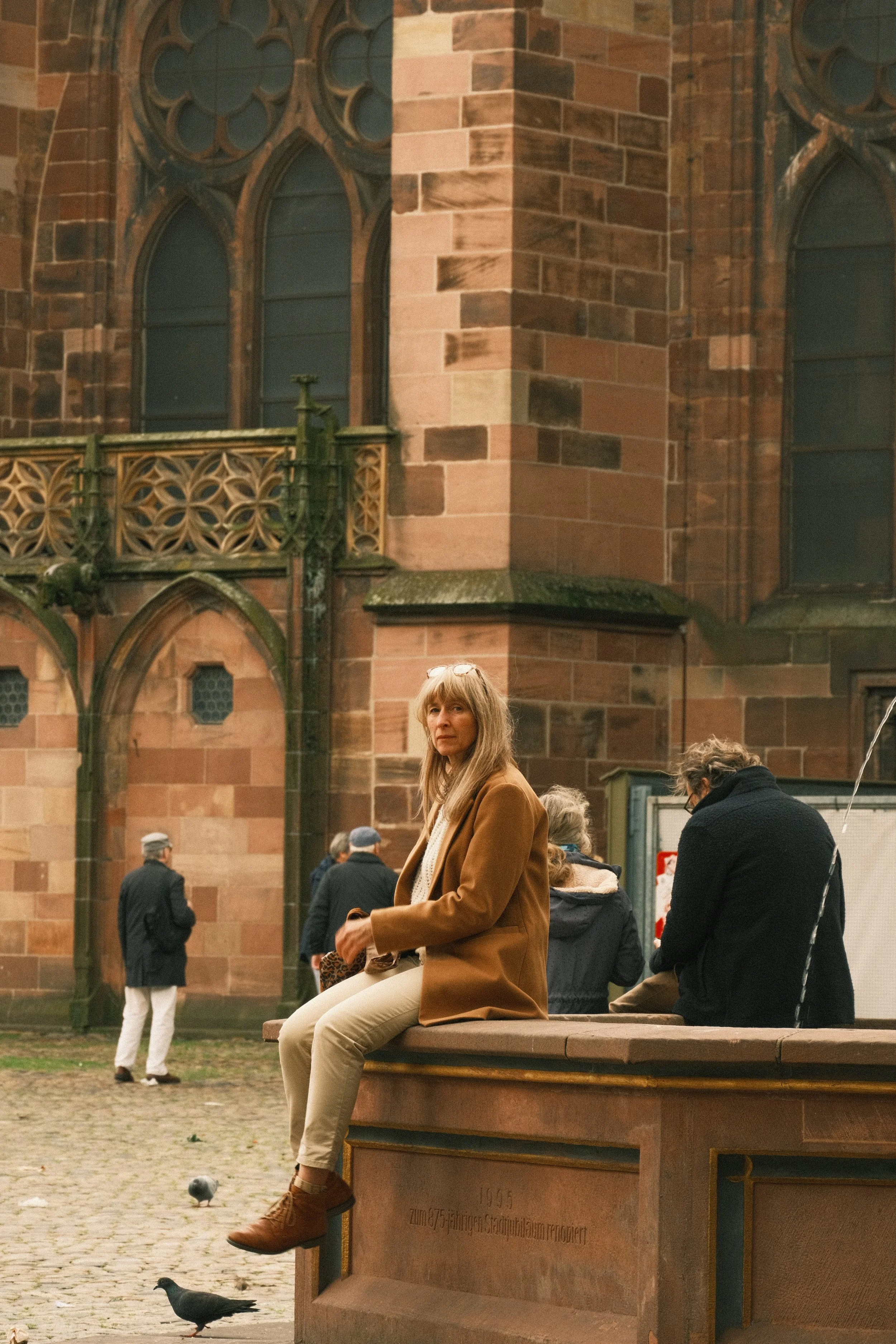 A woman with long blonde hair wearing a brown coat and beige pants is sitting on a stone bench in front of a historical red sandstone building with large arched windows. Several people are walking and standing around the area, and pigeons are on the ground in front of her.