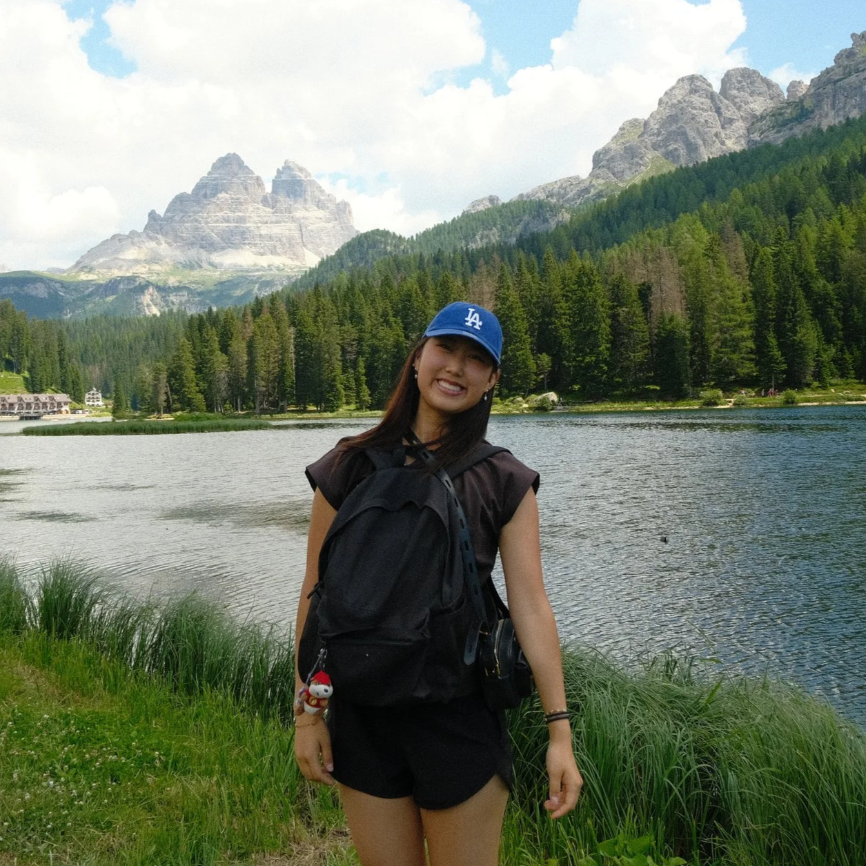 A woman smiling at camera standing by a lake with green trees and mountains in the background, wearing a blue Los Angeles Dodgers cap, black shirt, and carrying a black backpack.