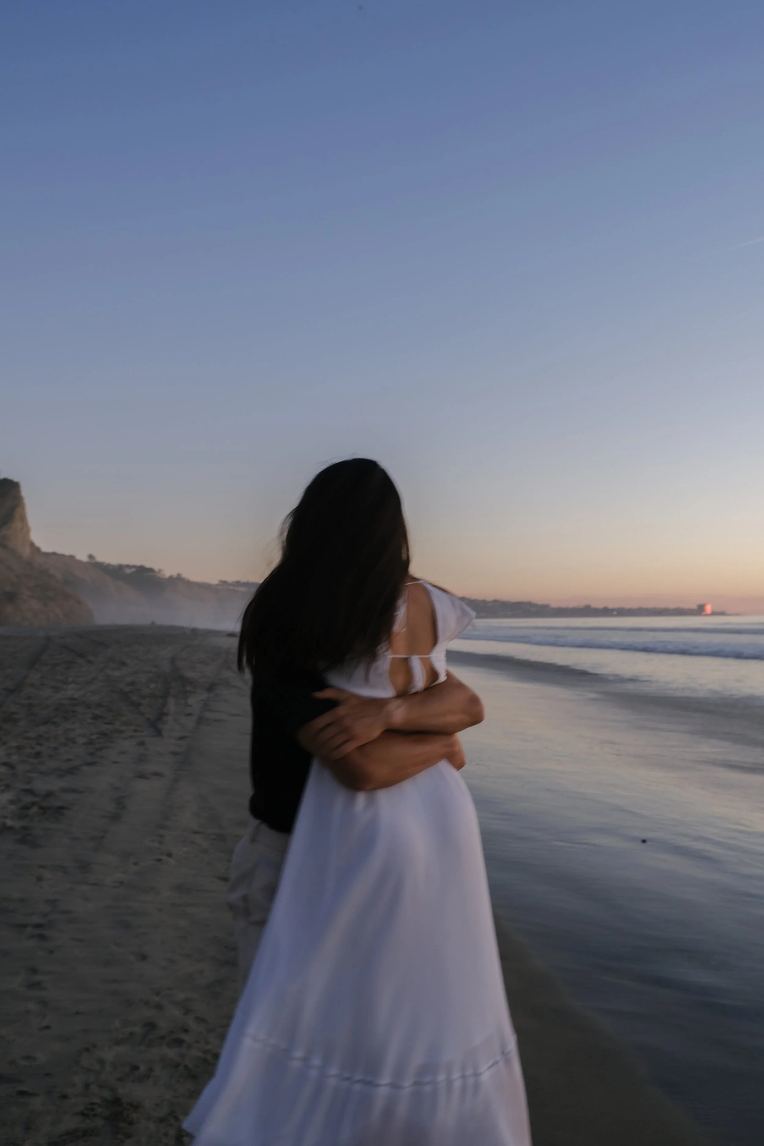 A woman in a white dress being embraced on a beach at sunset, with the ocean, cliffs, and a distant lighthouse in the background.