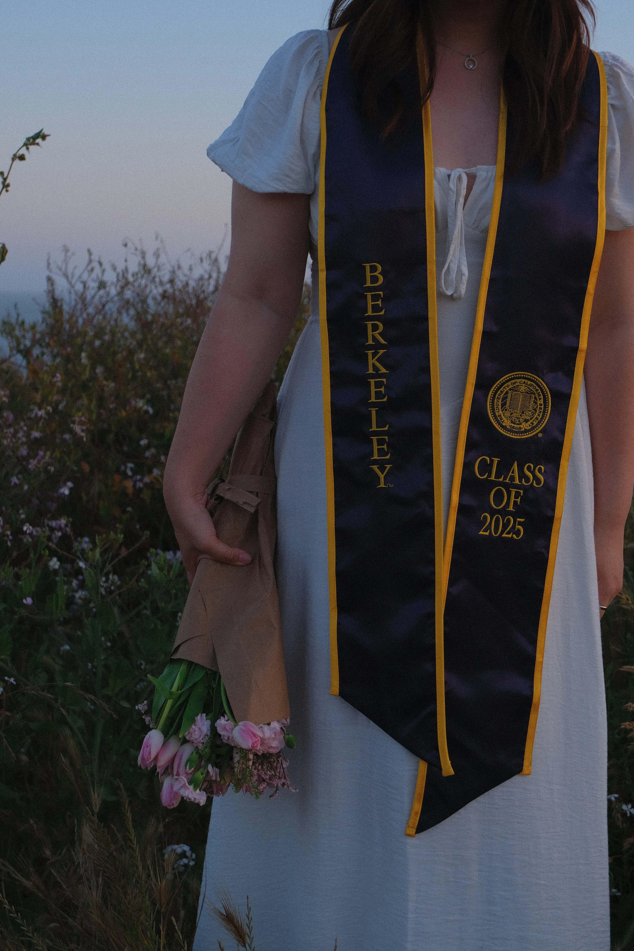 A person wearing a white dress with a graduation stole that reads 'Berkeley Class of 2025' and is holding a bouquet of pink flowers in front of greenery during sunset.