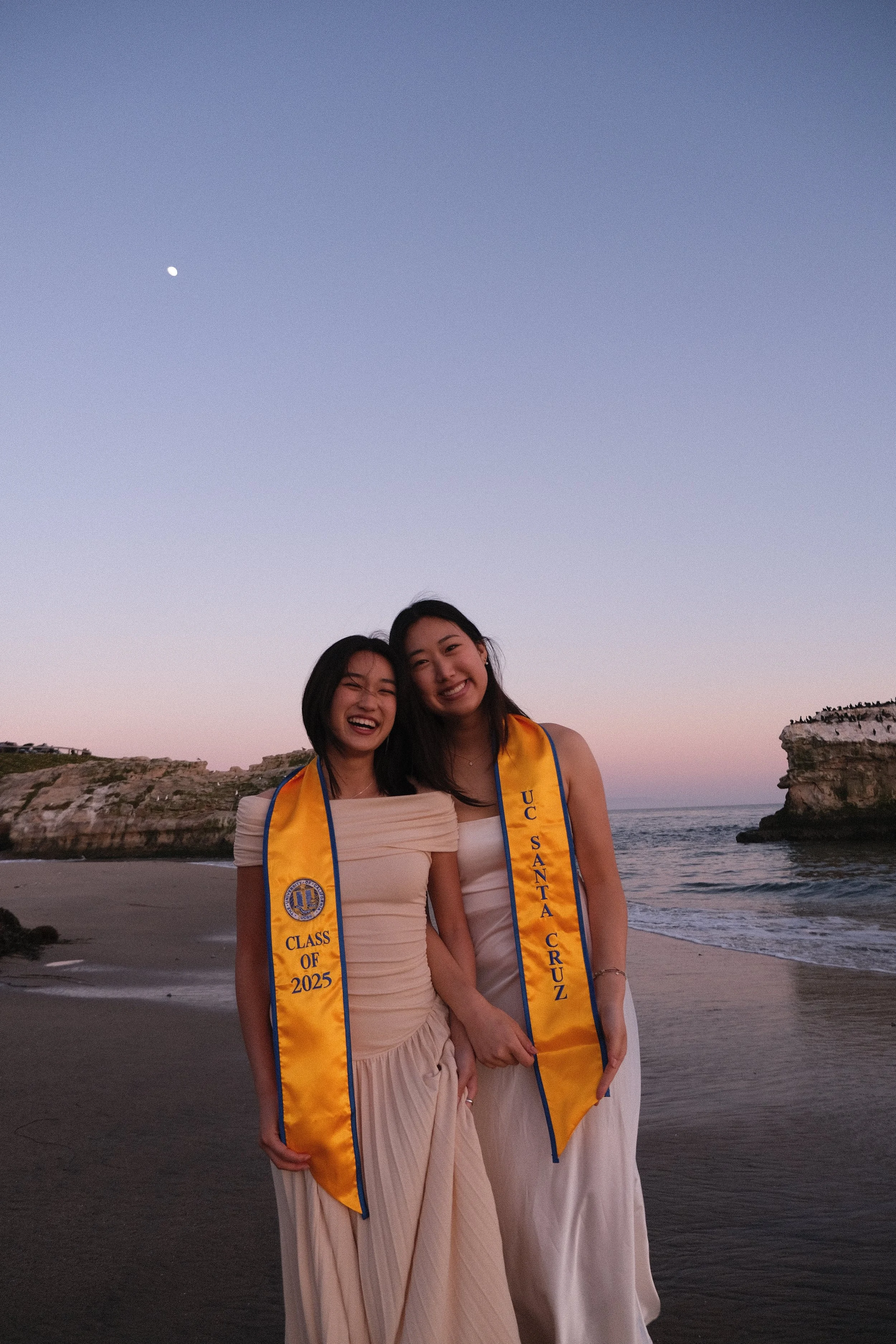 Two young women in formal dresses smiling on a beach at sunset, wearing yellow graduation stoles that read 'Class of 2025' and 'U.C. Santa Cruz'.