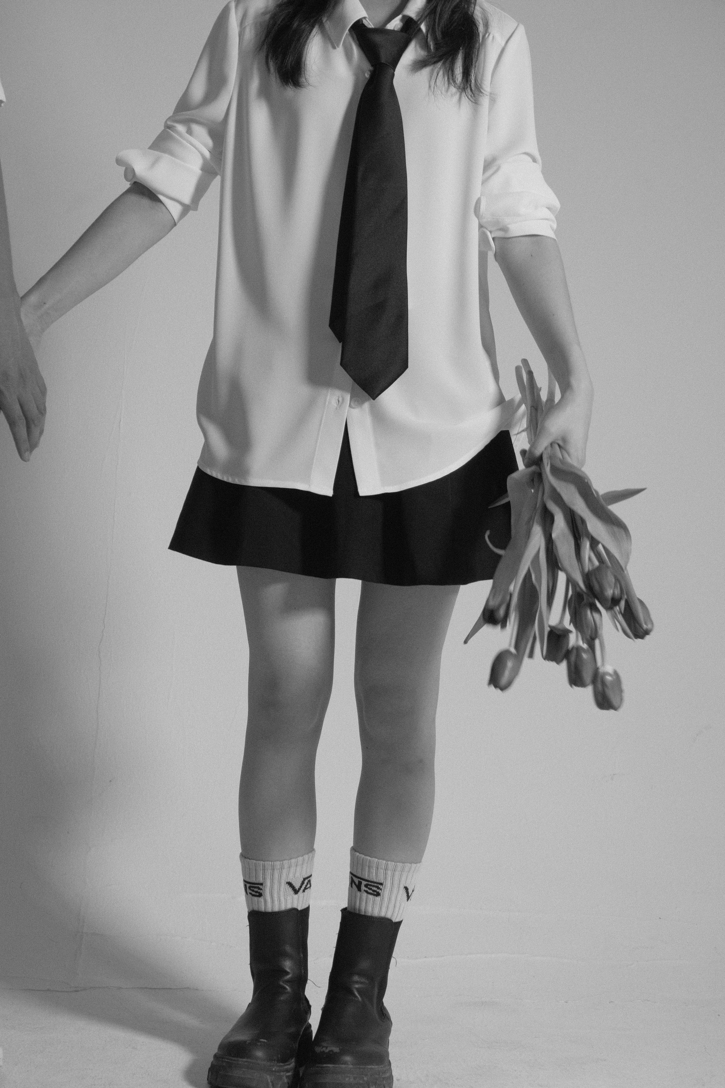 A person wearing a white shirt with rolled-up sleeves, a black tie, a black skirt, knee-high socks with writing, and black boots, holding a bunch of tulips.