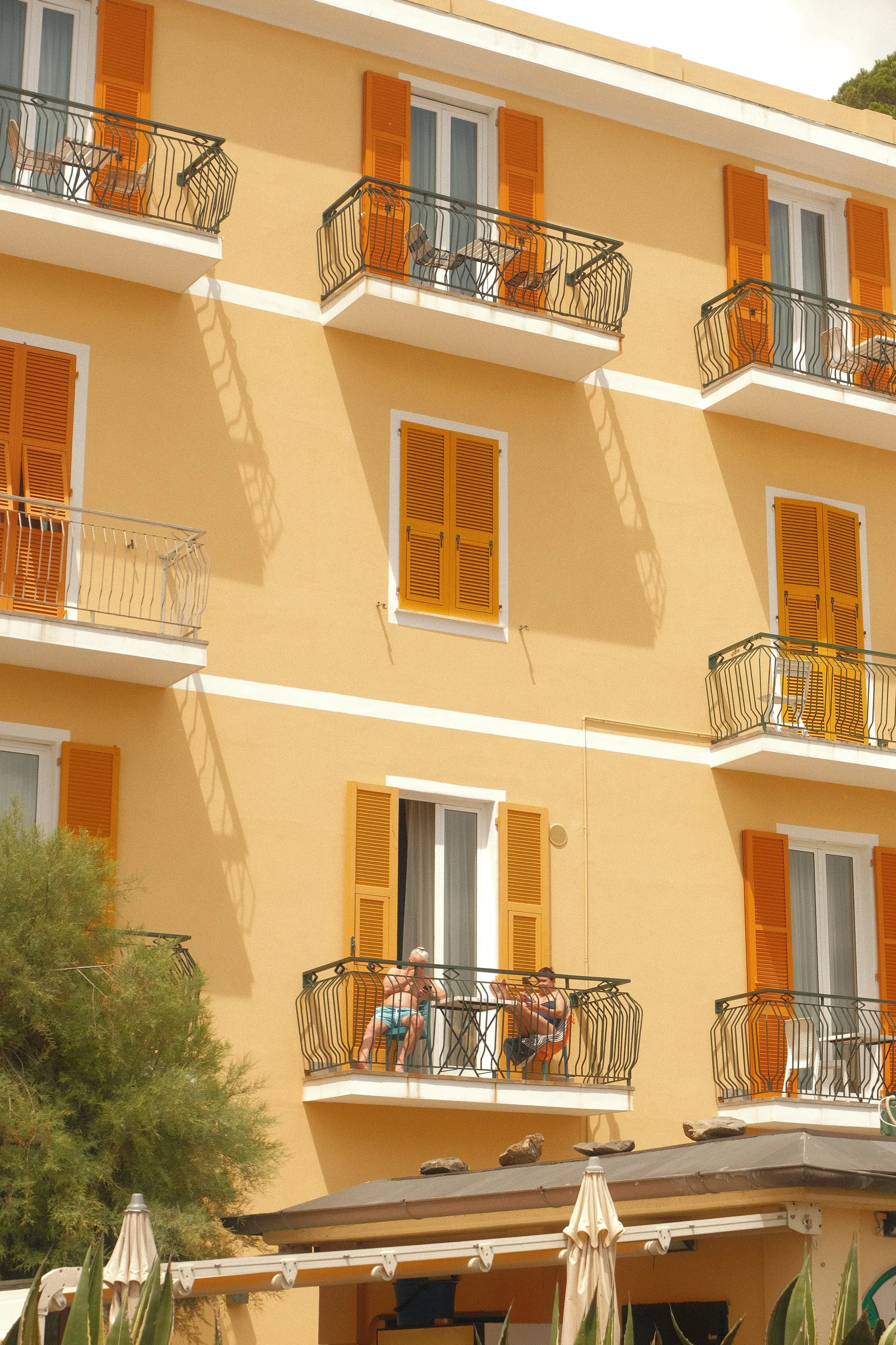 A yellow apartment building with multiple balconies featuring orange shutters, some with chairs and tables. Two people are sitting on a balcony on the second floor, enjoying the outdoors. The building has white trim around windows, and there are umbrellas and greenery at the bottom.