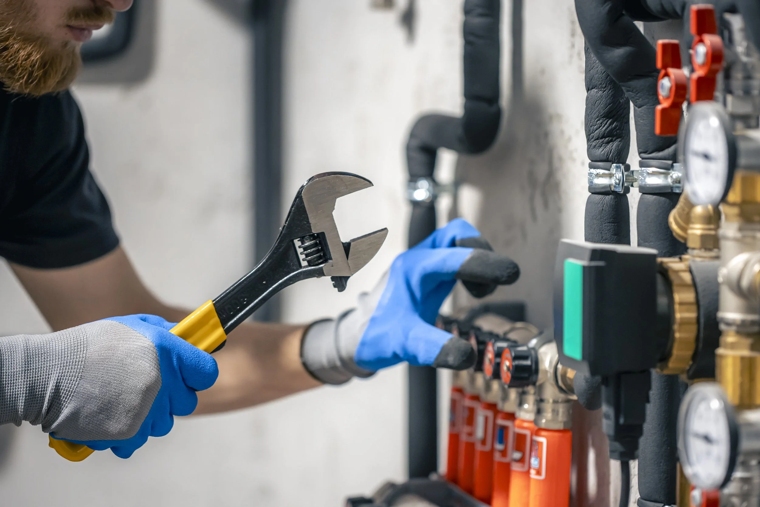 A person wearing blue gloves uses a wrench to work on pipes and gauges mounted on a wall.