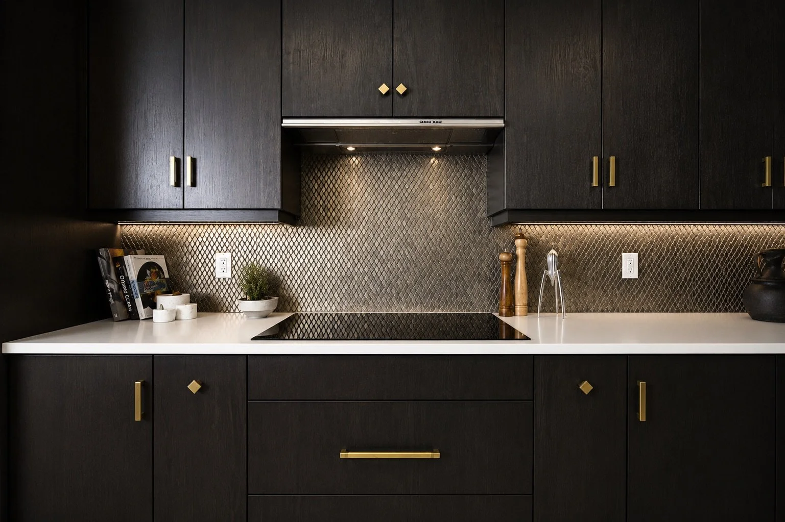 Modern kitchen with black cabinets, white countertop, gold handles, diamond-patterned metallic tile backsplash, built-in stovetop, and small decorations including a plant, salt and pepper shakers, books, and a black vase.