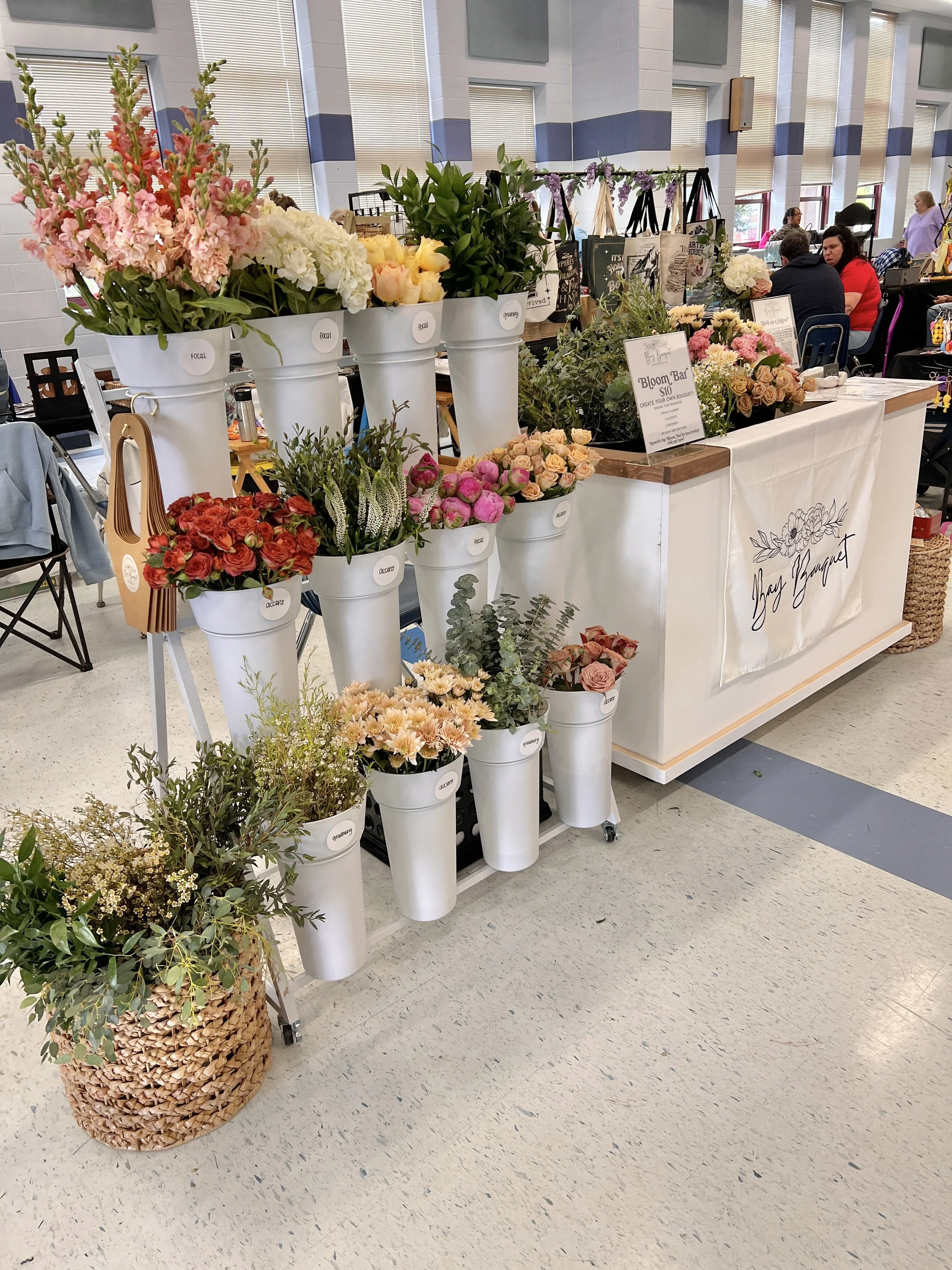 Display of various fresh flowers arranged in white buckets at an indoor market or craft fair, with people browsing and shopping in the background.