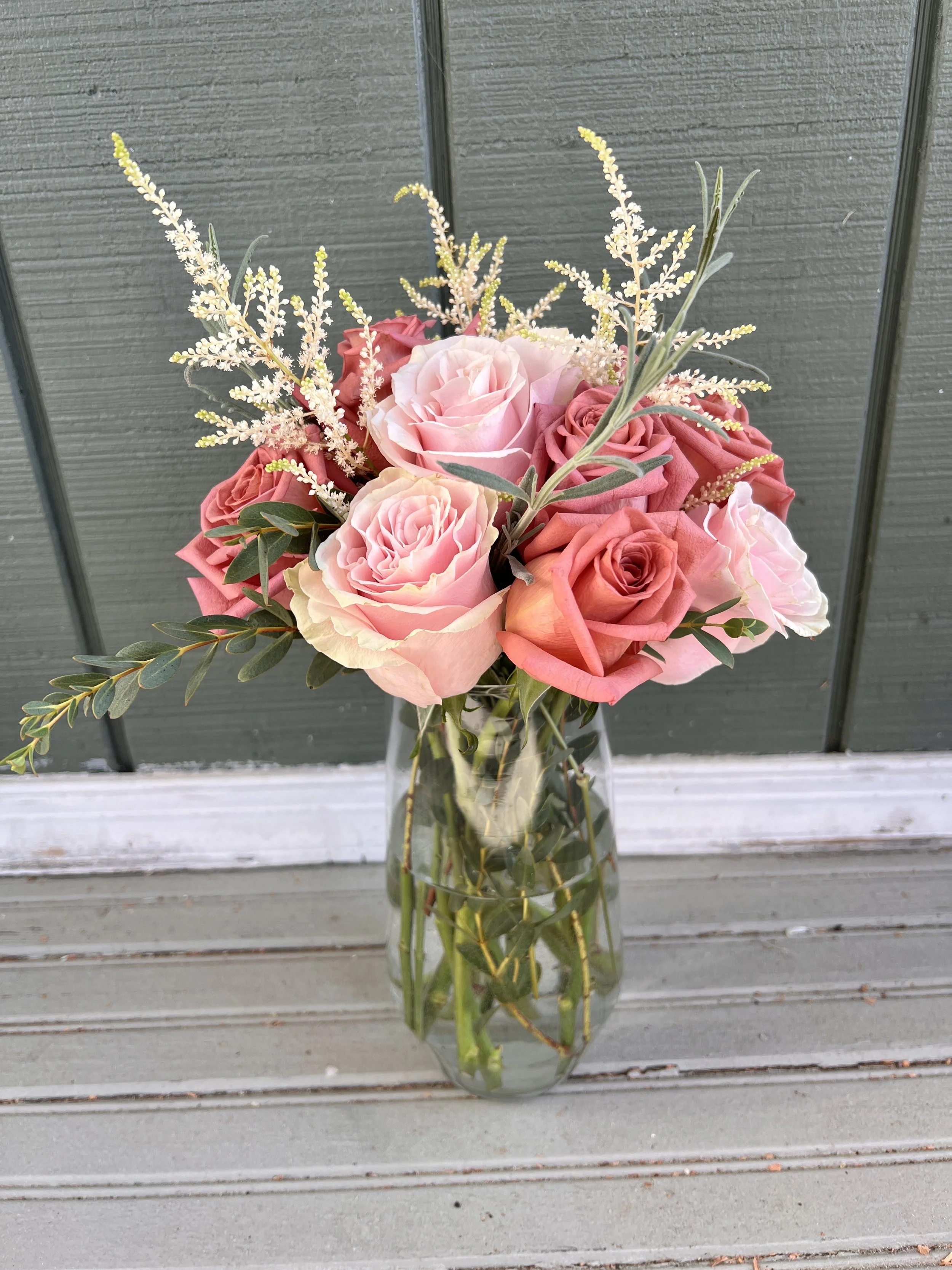 A bouquet of pink and red roses with green foliage and white accent flowers in a clear glass vase on a wooden surface.