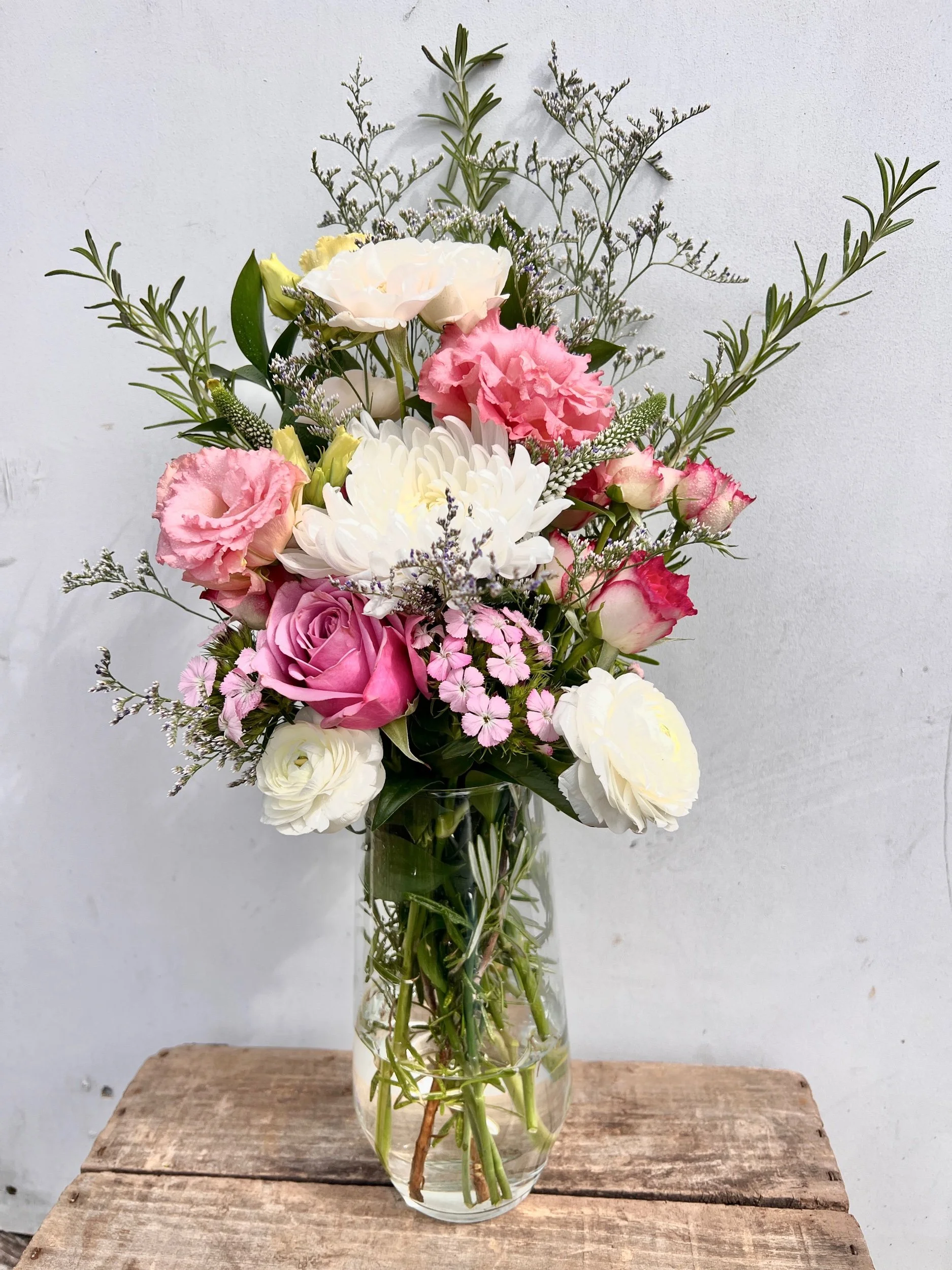 A glass vase filled with a variety of pink, white, and cream flowers, including roses, carnations, and chrysanthemums, with greenery and small filler flowers, placed on a rustic wooden surface against a plain white wall.