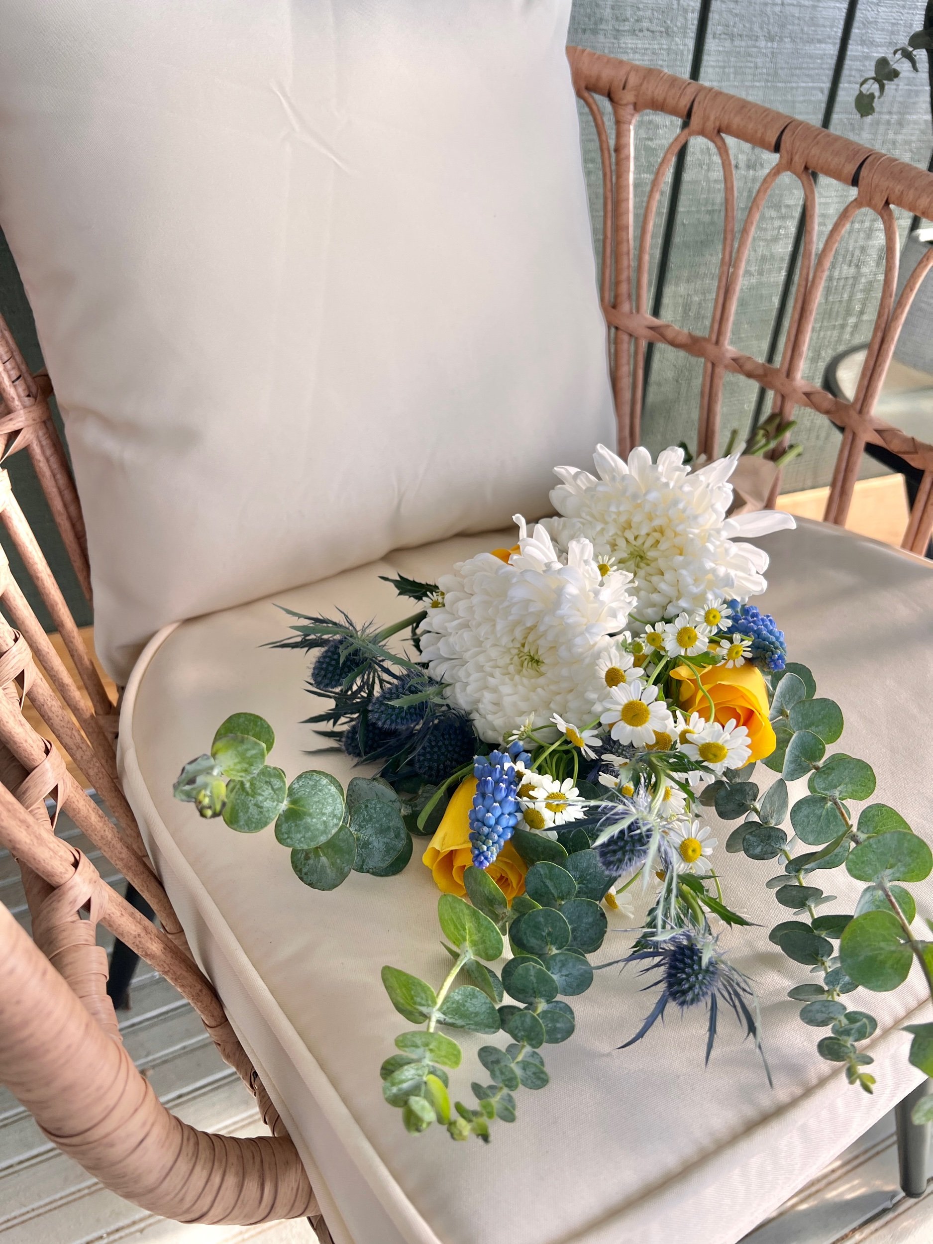 A bouquet of white daisies, blue thistles, yellow roses, and green foliage resting on a woven chair with a white cushion, next to a beige table.