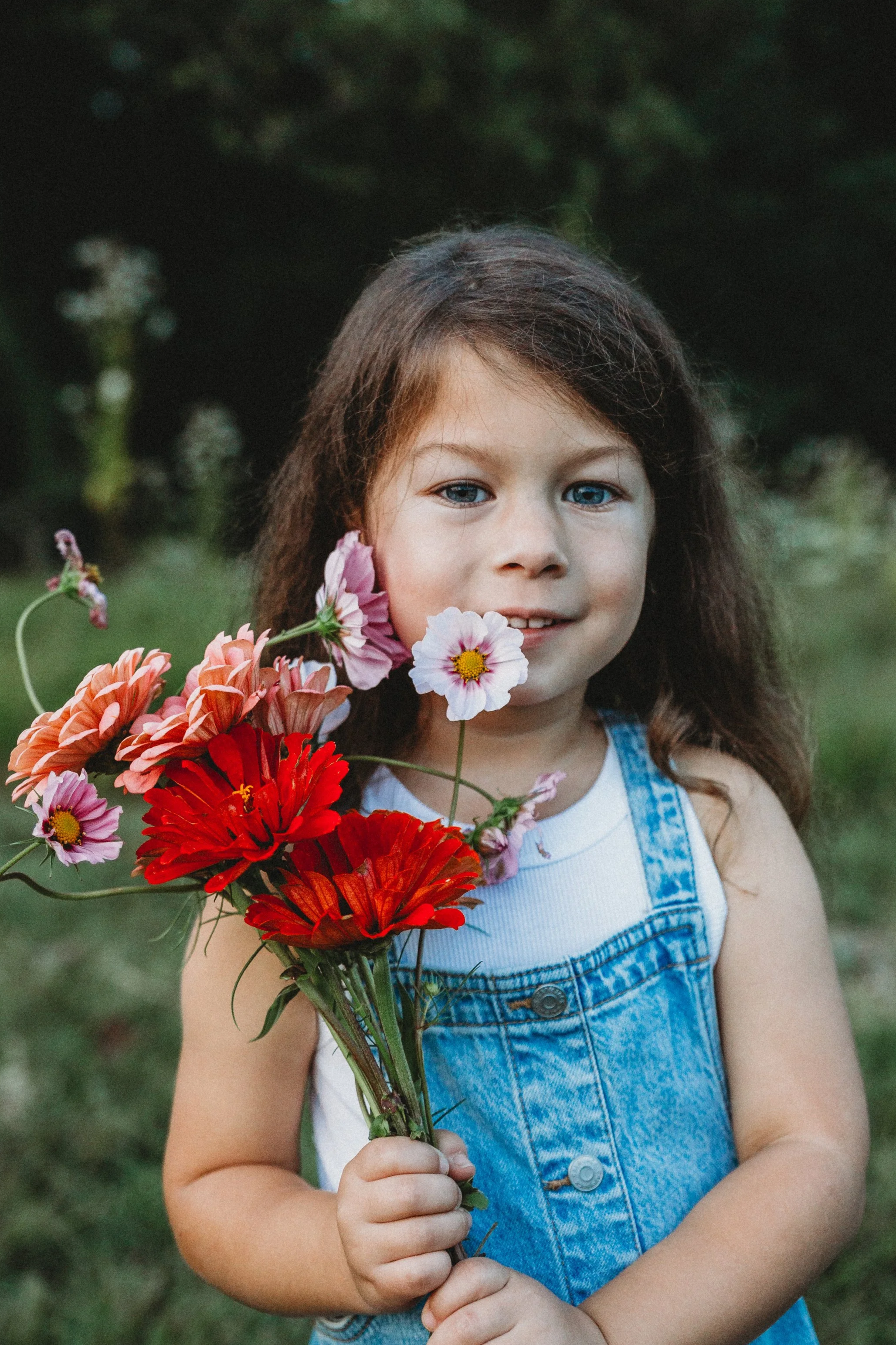 A young girl with brown hair and blue eyes holding a bouquet of colorful flowers including red, pink, and white blooms with yellow centers, outdoors with a blurred green background.