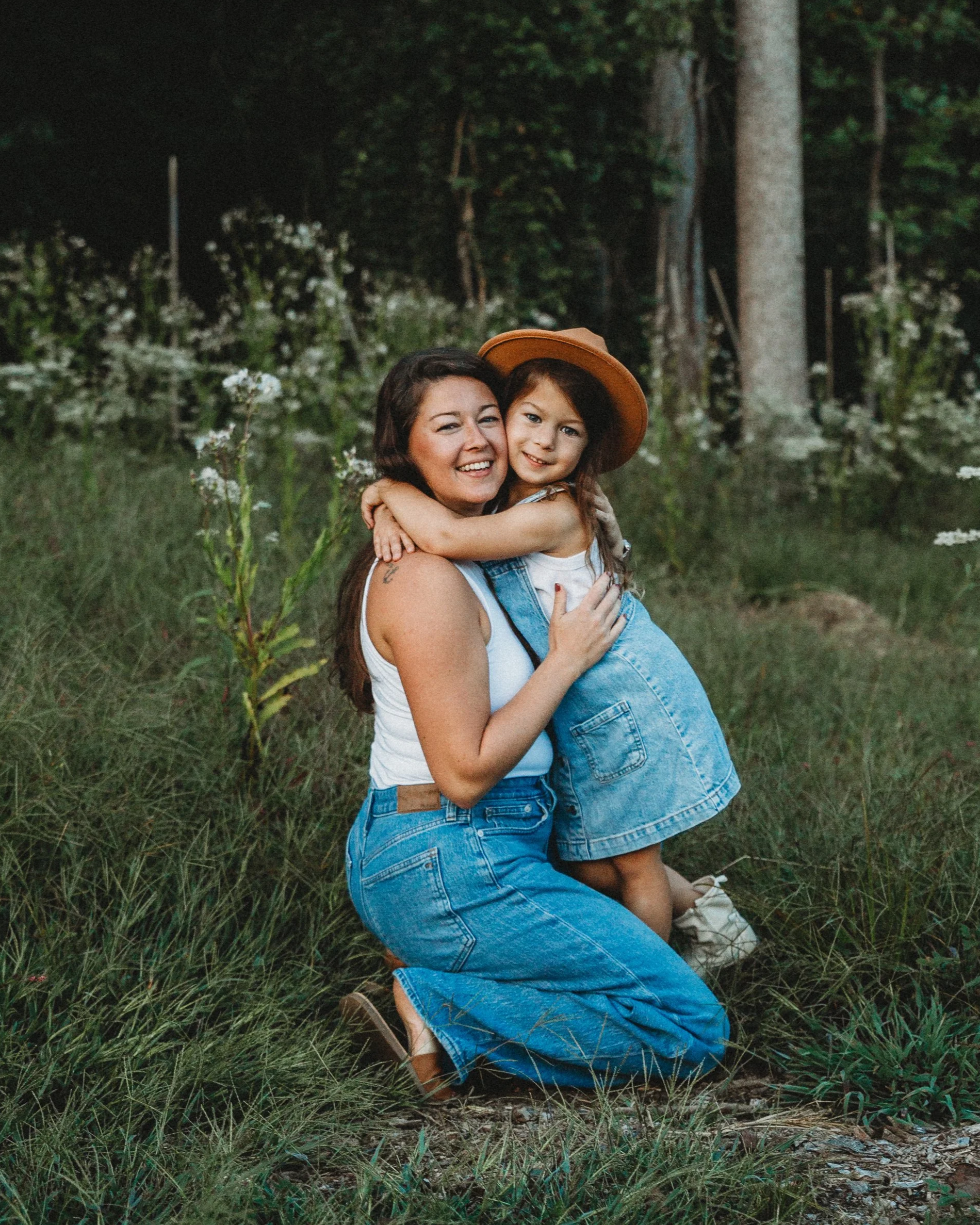 A woman and a young girl hugging in a forested area, both smiling. The woman has long dark hair and is wearing a white tank top and blue jeans. The girl has dark hair, is wearing a brown hat, white shirt, and blue overalls. They are surrounded by grass and white flowering plants.