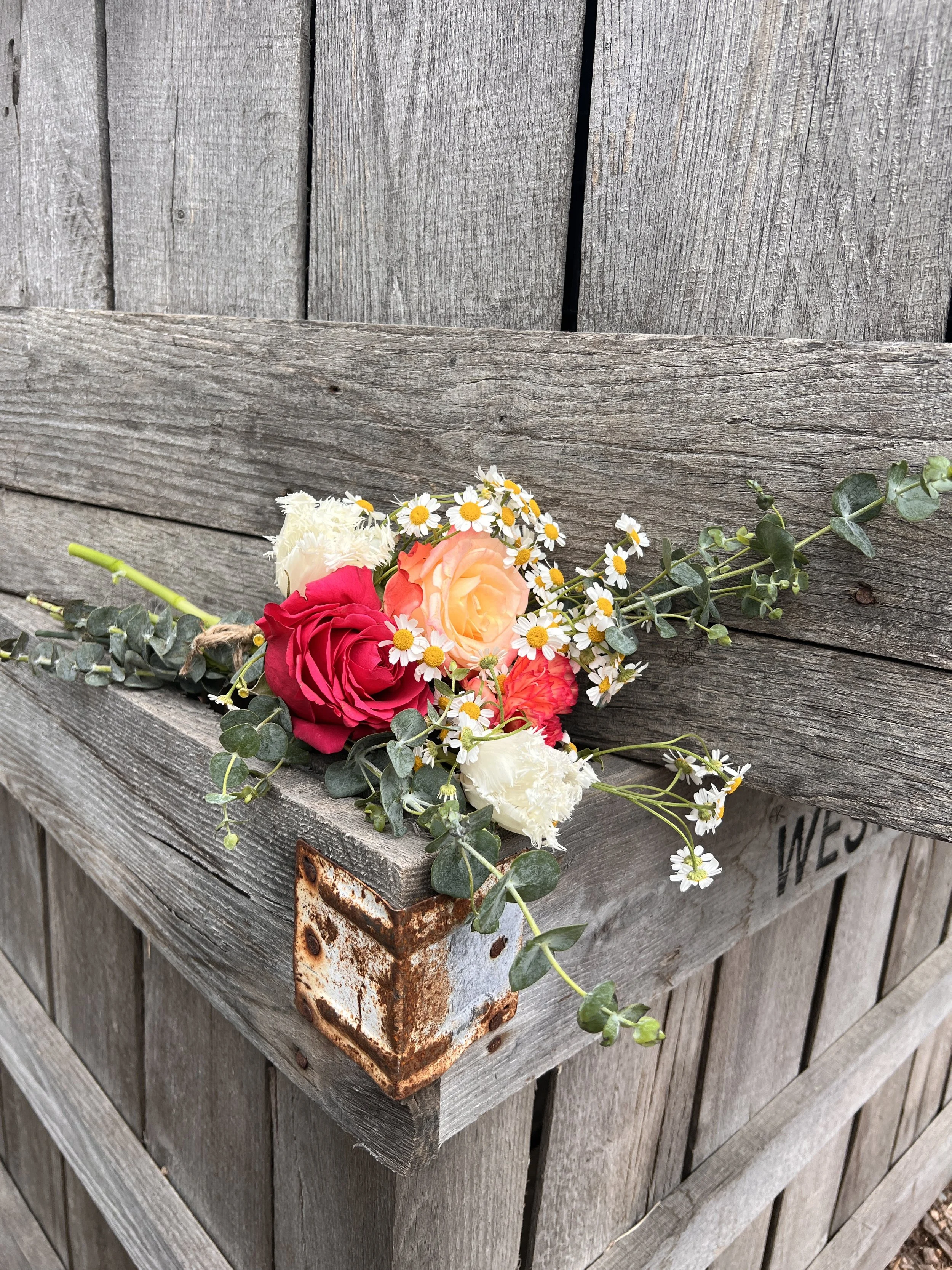 A bouquet of colorful flowers, including red, yellow, and white blooms, laid on a rustic wooden box attached to a weathered wooden fence.