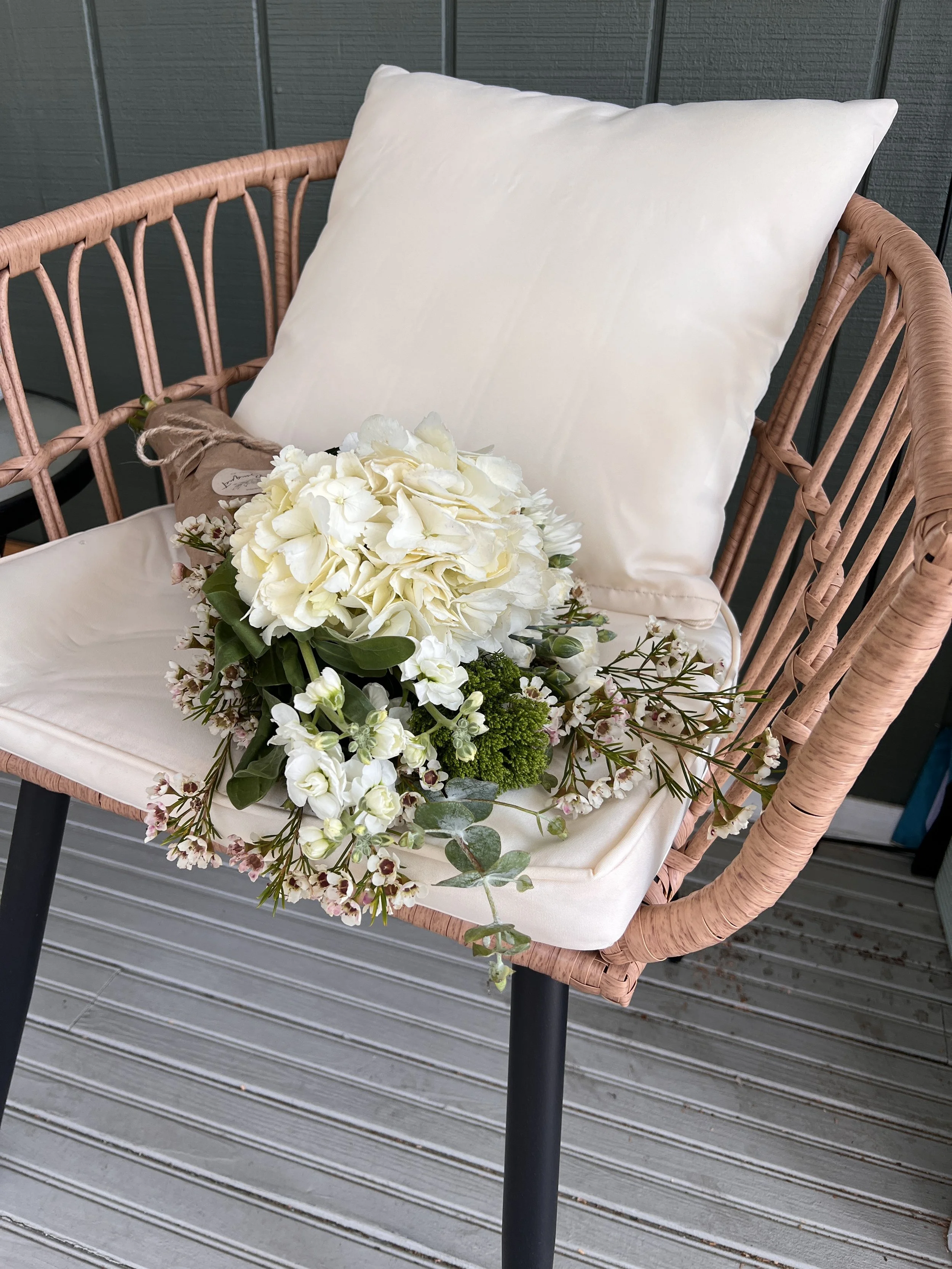 A pink wicker outdoor chair with a white cushion, decorated with a bouquet of white flowers and greenery, placed on a grey wooden deck.