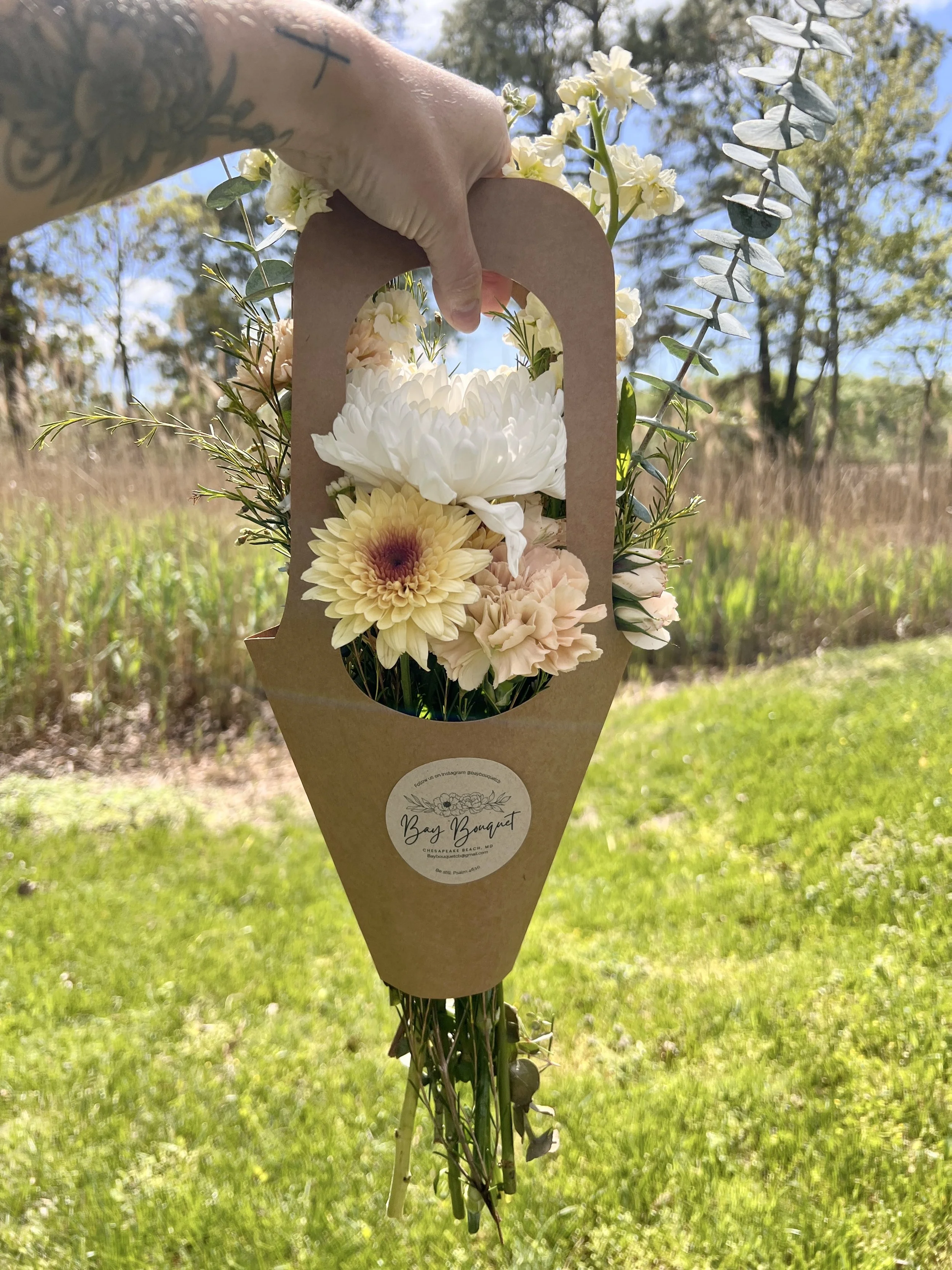 A bouquet of various flowers including white and pale yellow blooms, wrapped in brown paper with a label reading "Bay Bouquet," held outdoors against a background of blue sky, green trees, and grass.