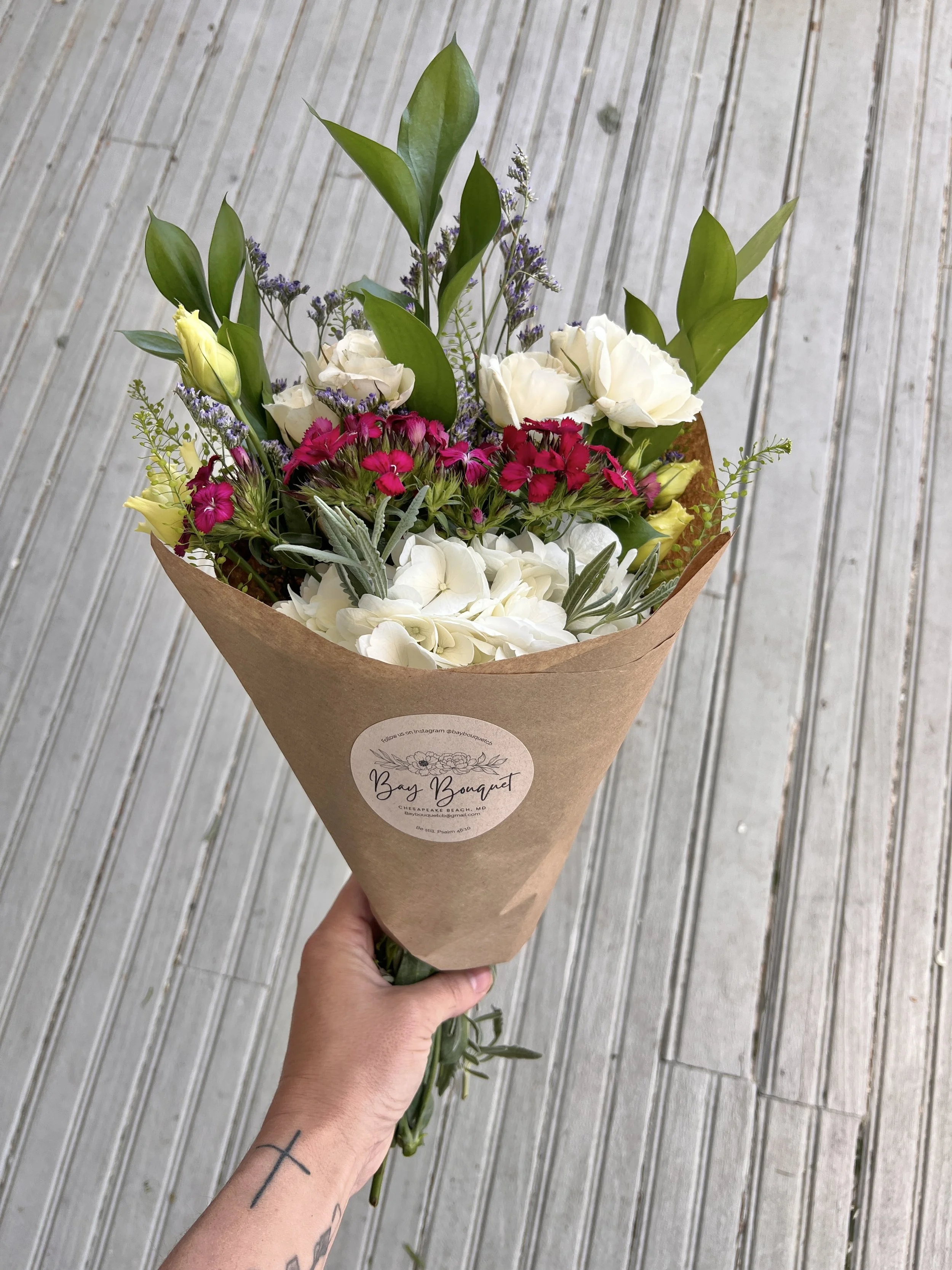 A person holding a bouquet of mixed flowers wrapped in brown paper, with a sticker that reads "Bay Bouquet," against a background of wooden floorboards.
