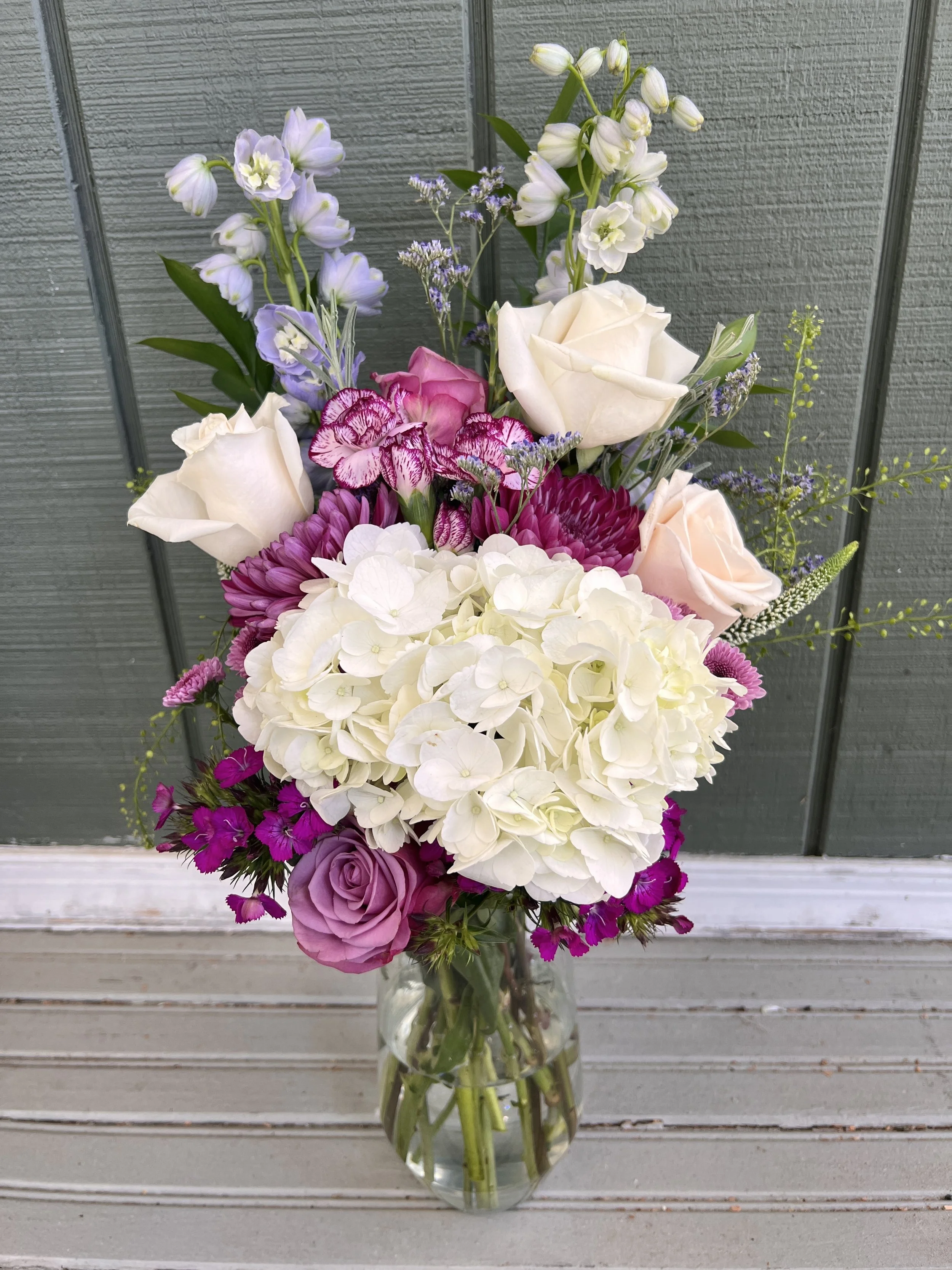 A floral arrangement in a glass vase with white roses, purple and pink carnations, hydrangeas, and various filler flowers against a wooden background.
