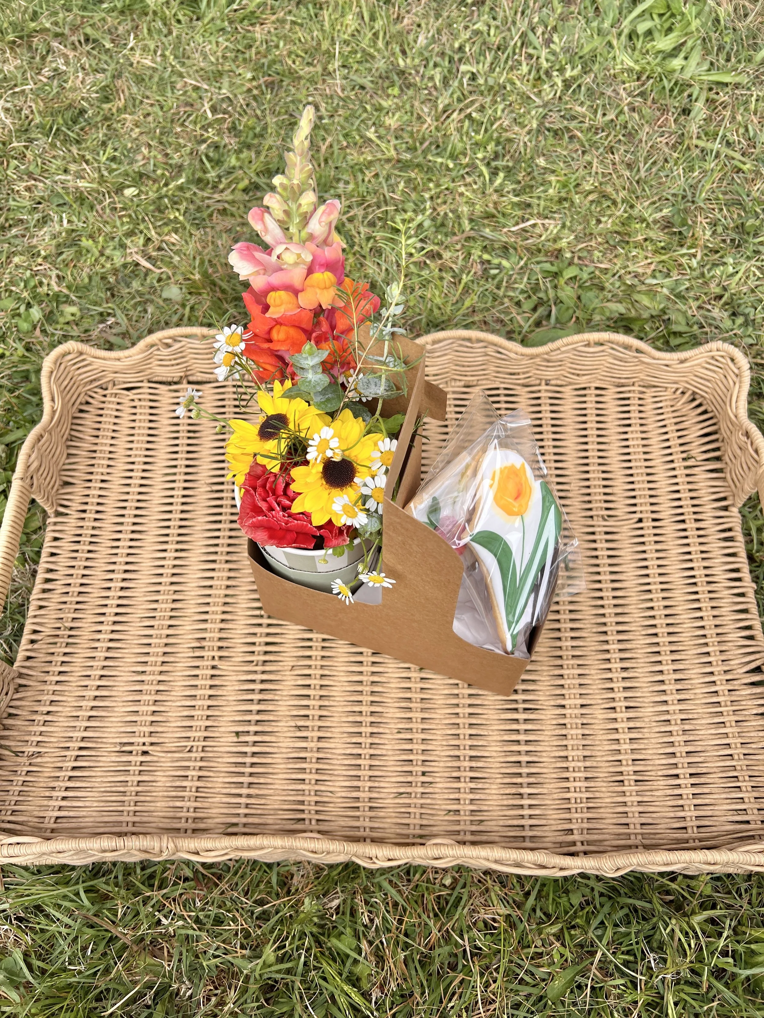 A wicker basket with a bouquet of various colorful flowers and a wrapped gift on a grassy surface.