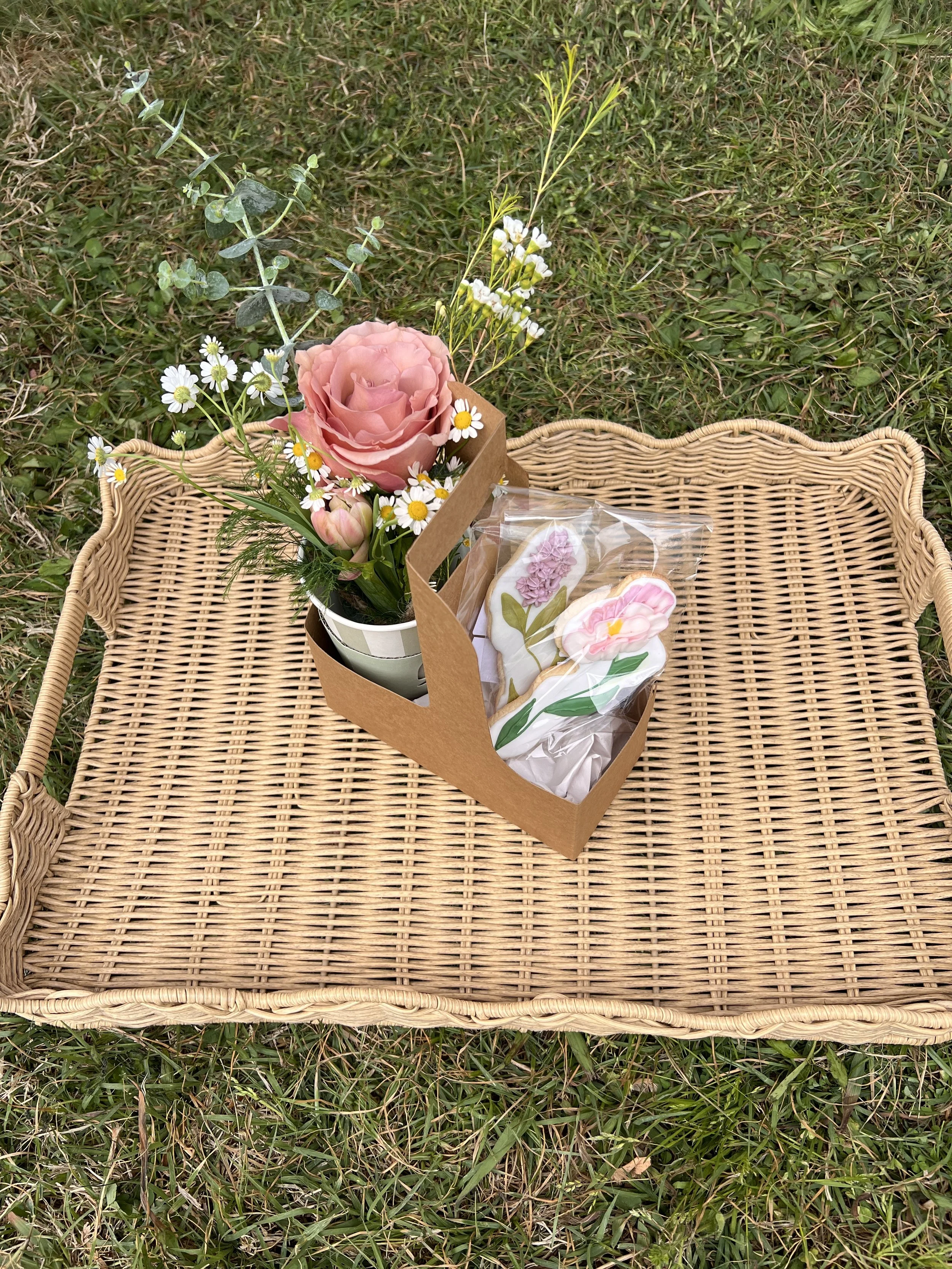 A wicker basket on grass containing a pink rose, daisies, and other flowers, with decorated cookies wrapped in clear plastic.