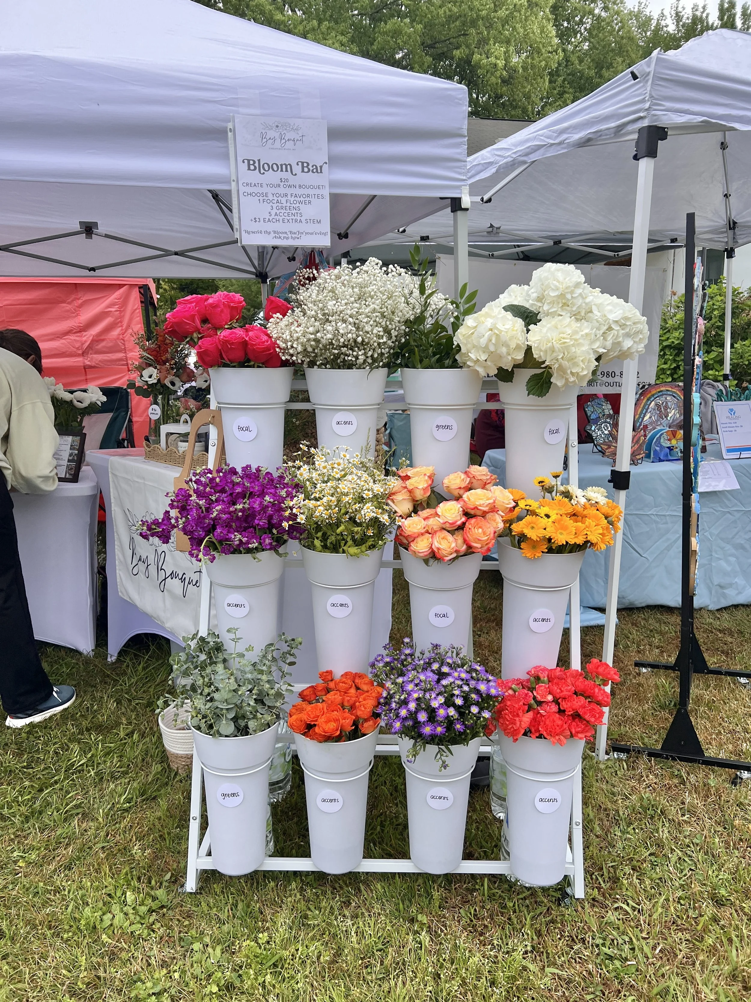 A flower stand at an outdoor market with white buckets of colorful flowers including roses, hydrangeas, daisies, and other blooms. White tents and a sign above advertise a 'Bloom Bar' for creating custom bouquets, with options for focal flowers, gree