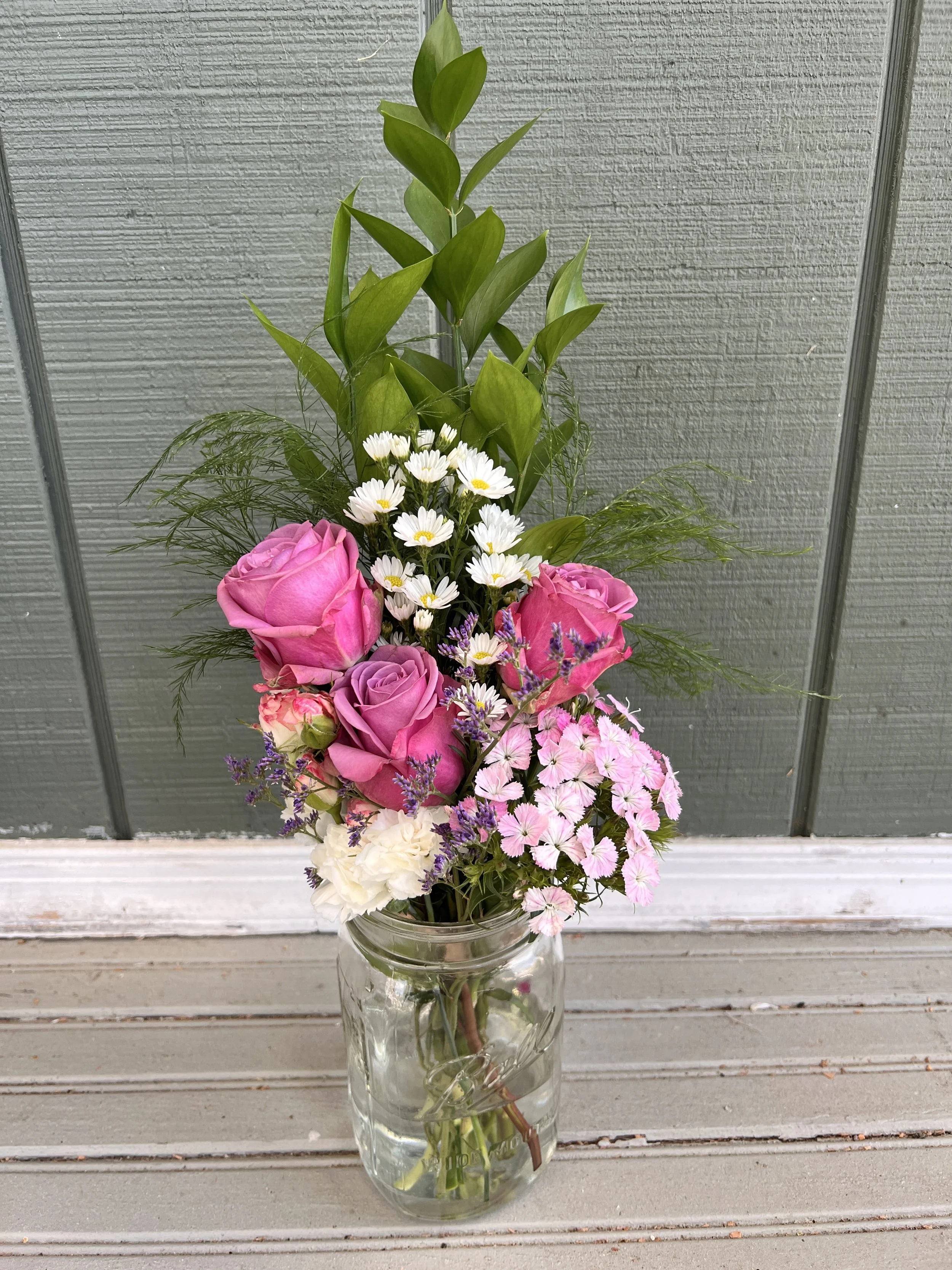 A tall glass vase with a bouquet of mixed pink, white, and purple flowers, including roses, daisies, and phlox, placed on a light gray wooden surface against a gray textured wall.