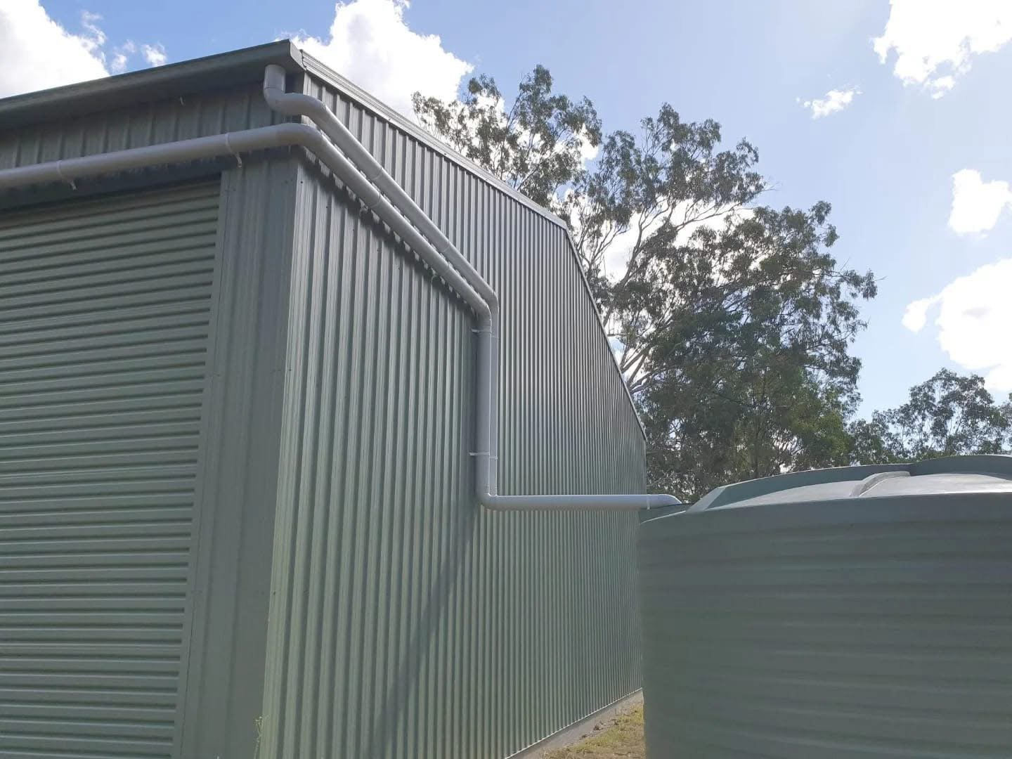 Side view of a large metal industrial building with a corrugated metal exterior, featuring an elevated window and white drainage pipes on the wall, with trees and blue sky in the background.