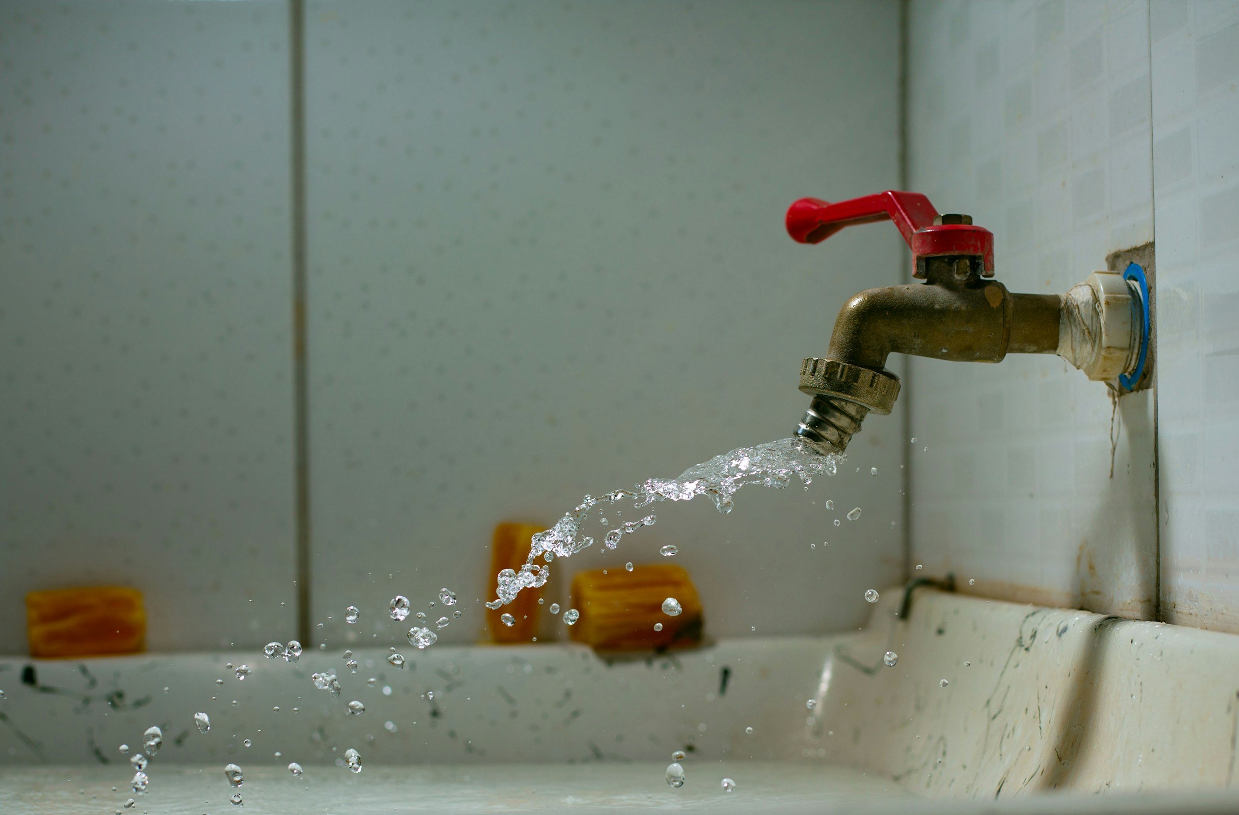 A water faucet with a red handle pouring water into a sink, with orange sponges on the sink's edge.