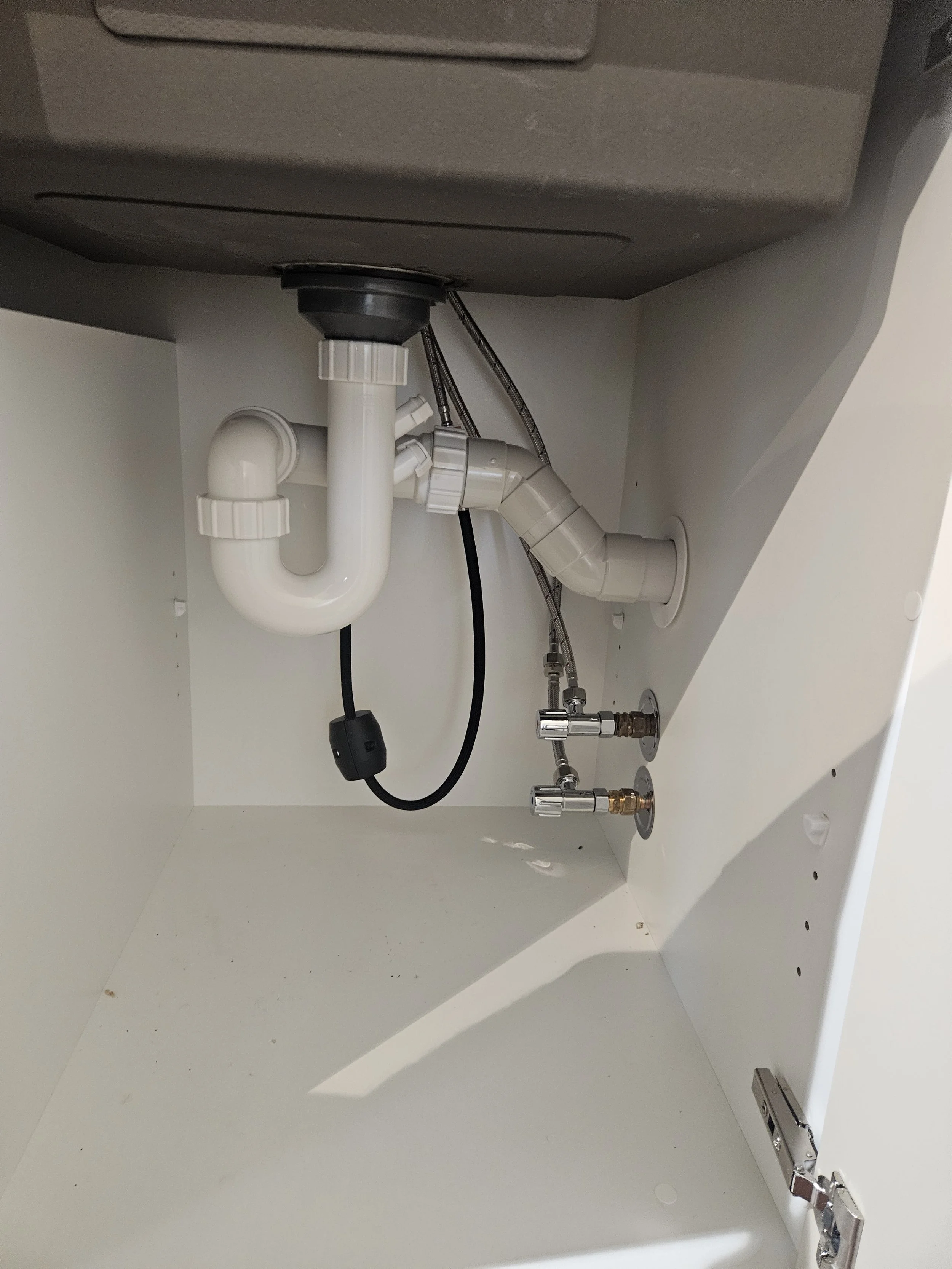 Under sink plumbing with white PVC pipes, shut-off valves, and chrome fixtures inside a cabinet.