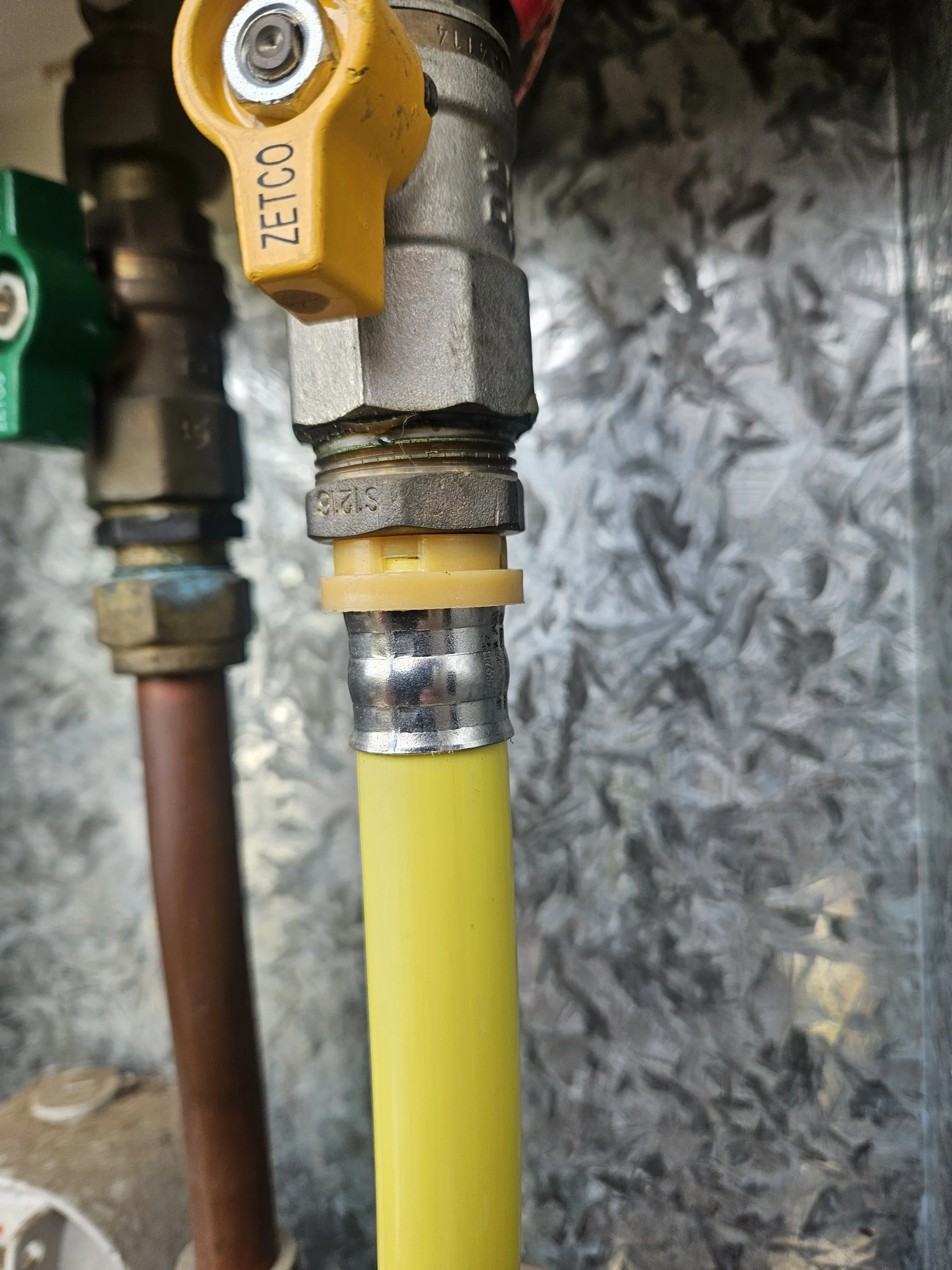 Close-up of a yellow gas shut-off valve connected to a yellow pipe, with a metallic fitting and a black control knob, against a textured metal background.