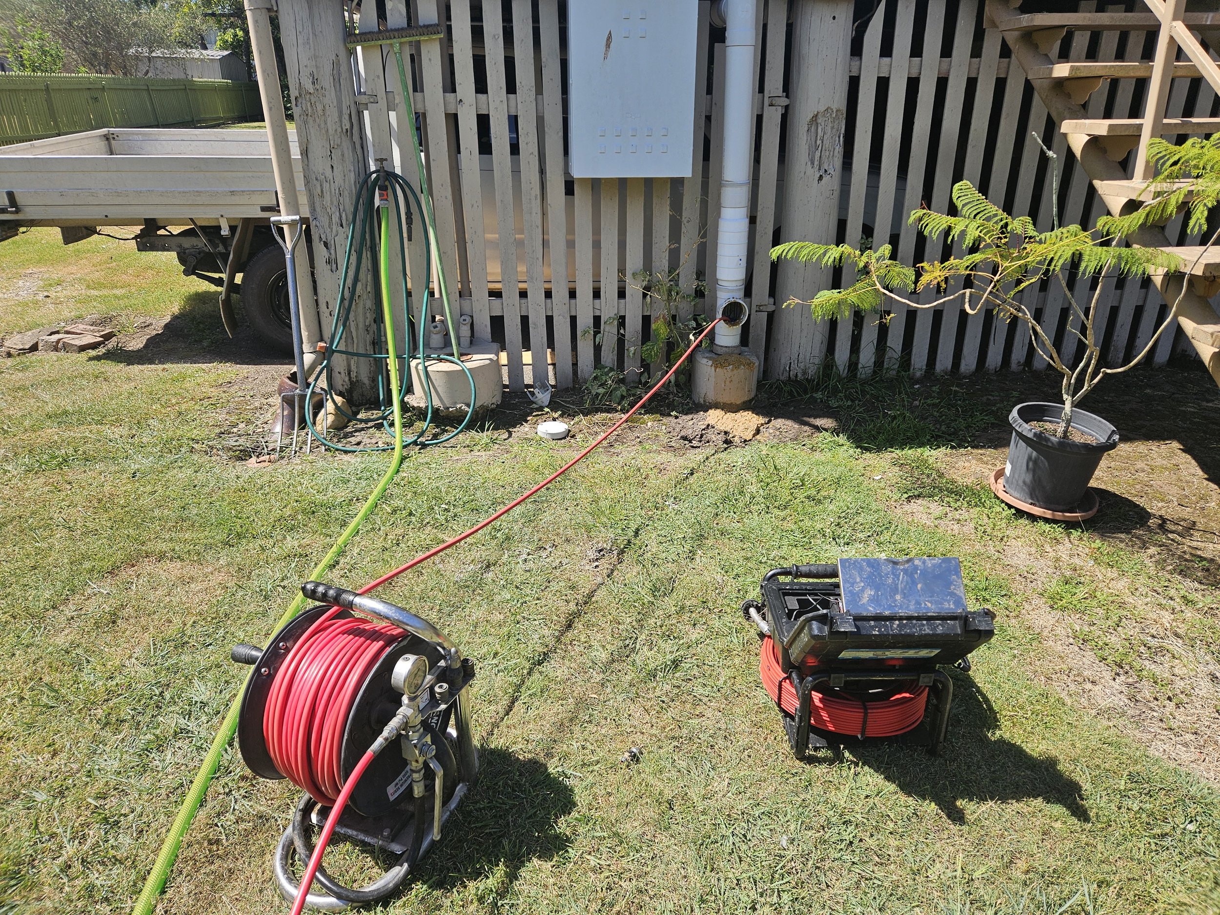 Tools and equipment for outdoor electrical work including a reel of red wire, a black toolbox, a green hose, and a yellow extension cord, set up on a grassy area near a wooden fence and stairs.