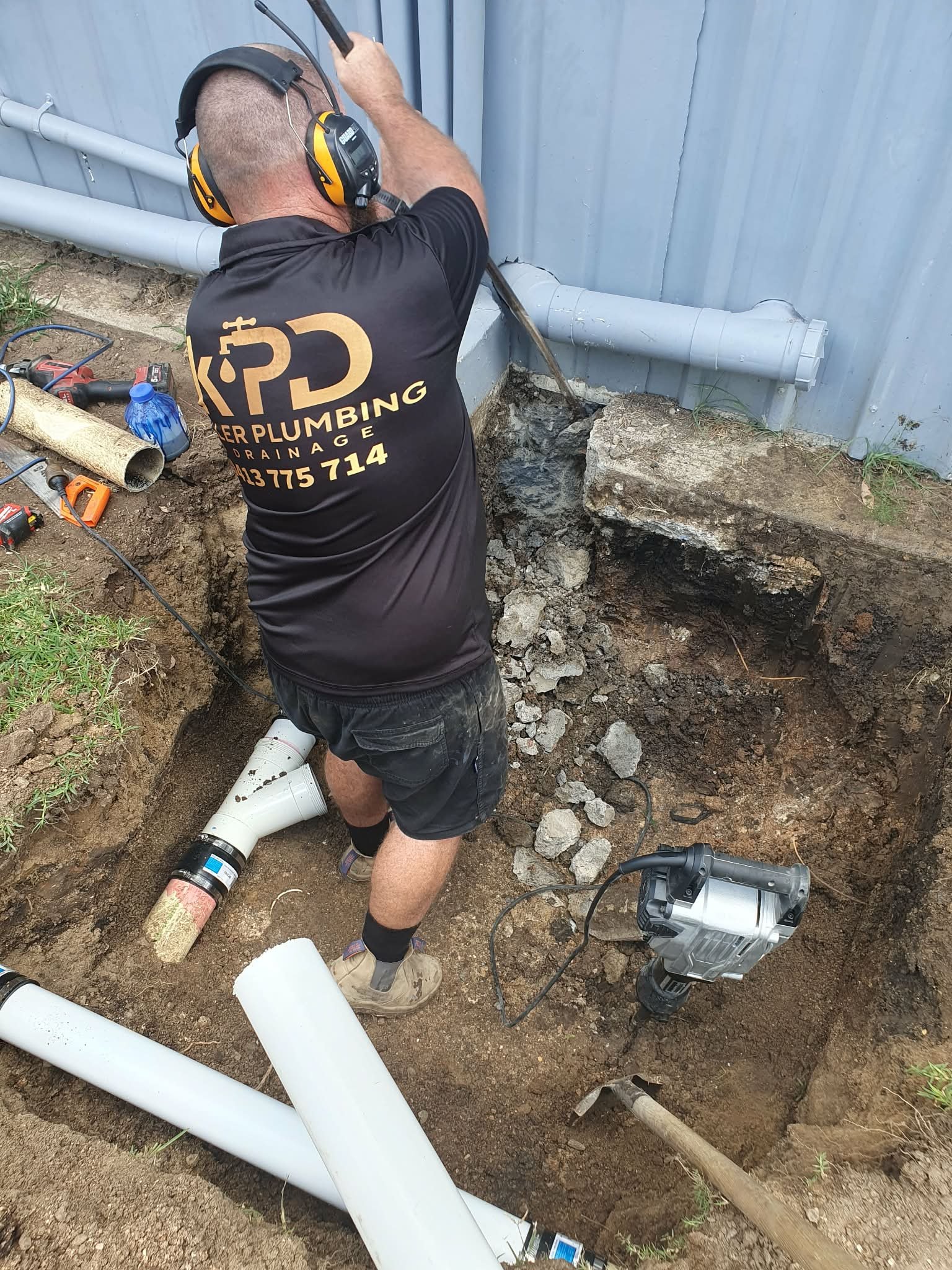 A plumber installing or repairing underground pipes outside a building, using power tools and standing in a trench with dug-up dirt and rocks.